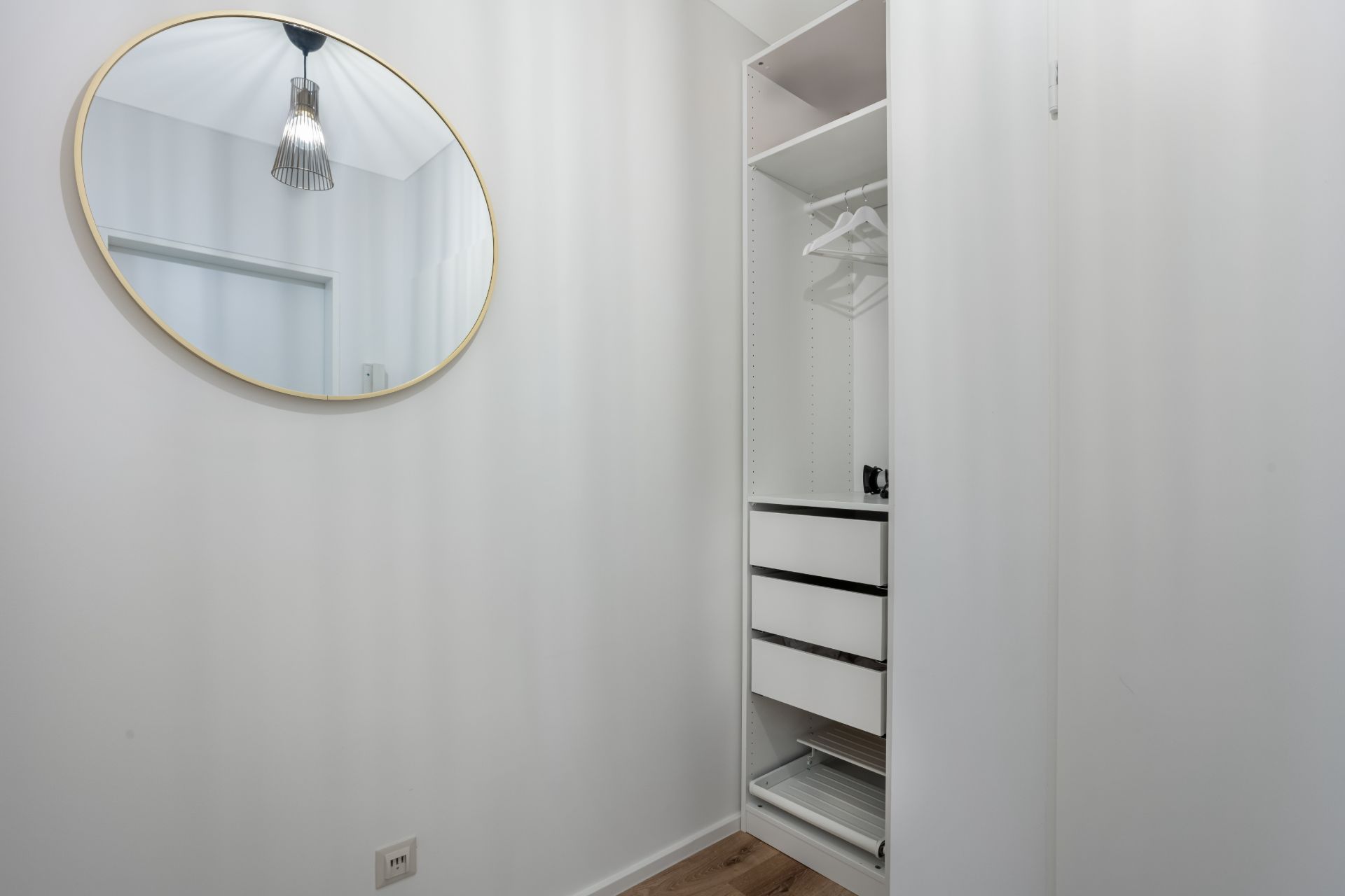 Offener Kleiderschrank mit Schubladen und Spiegel an einer weißen Wand.