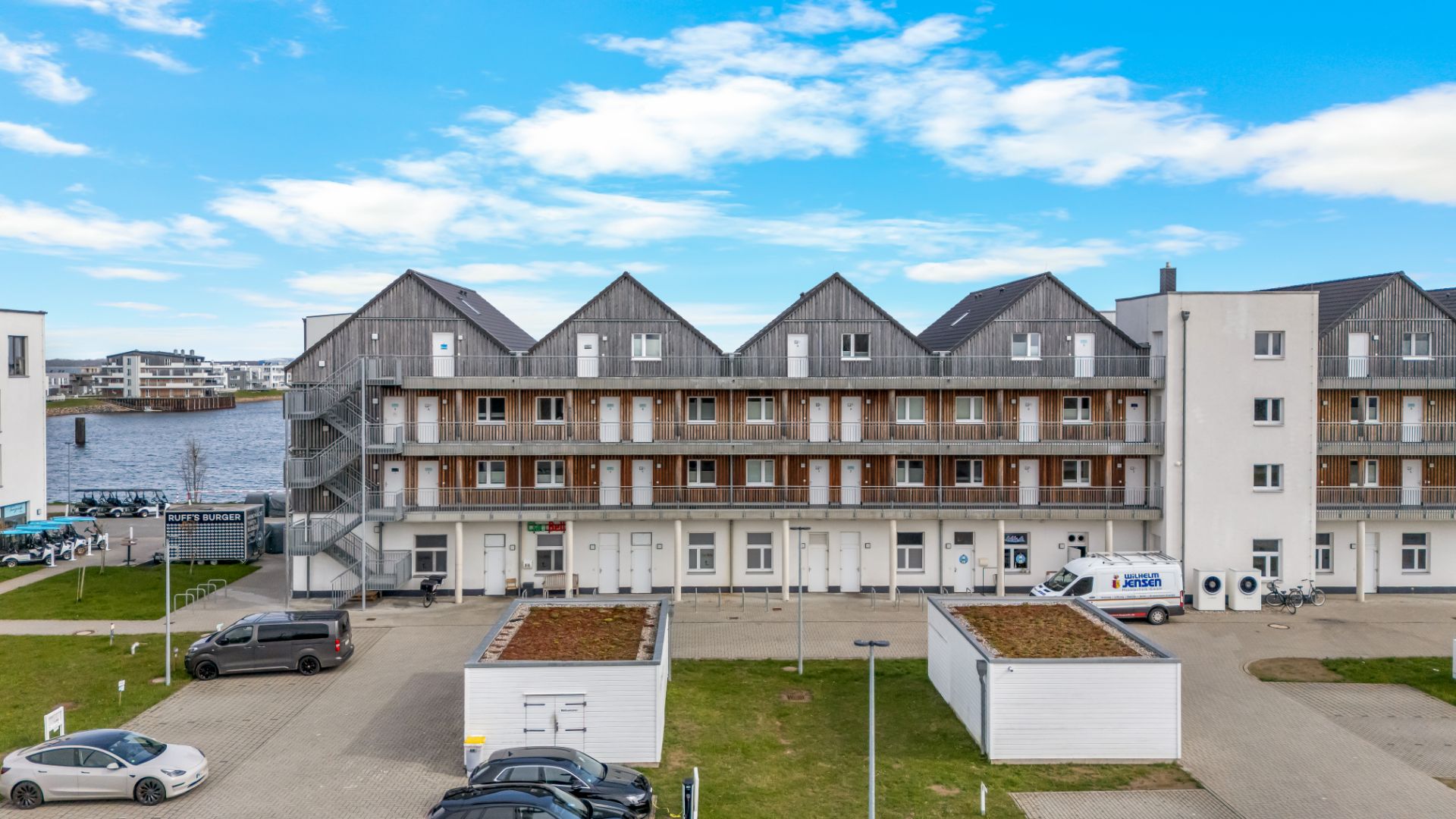Wohngebäude mit Holzbalkonen, mehreren Eingängen und Autos auf dem Parkplatz, im Hintergrund Wasser.