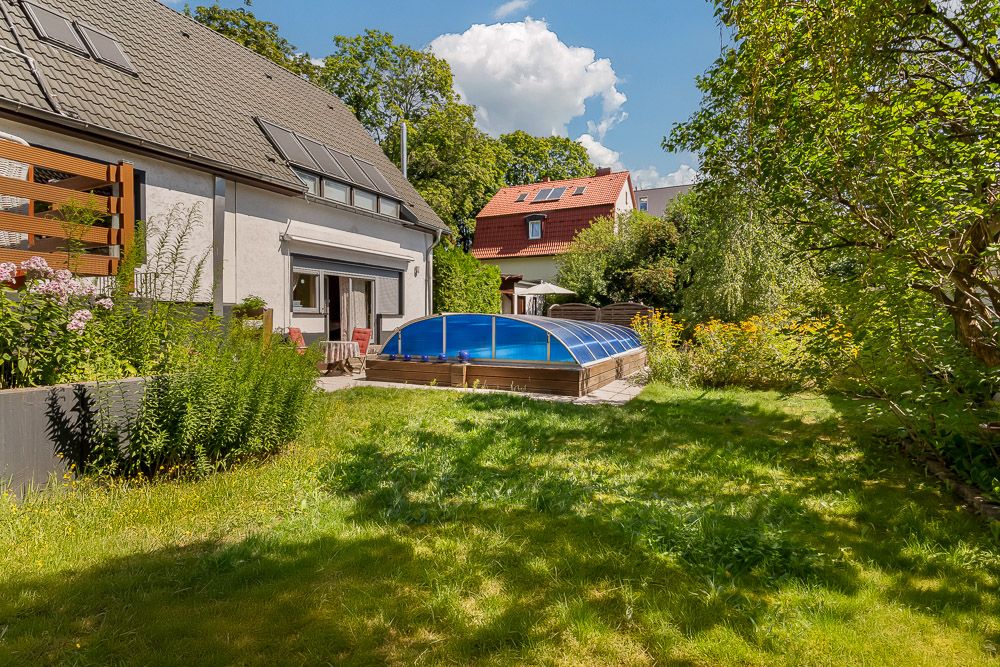 Garten mit überdachtem Pool, Rasenfläche, Blumenbeeten und zwei Wohnhäusern im Hintergrund.