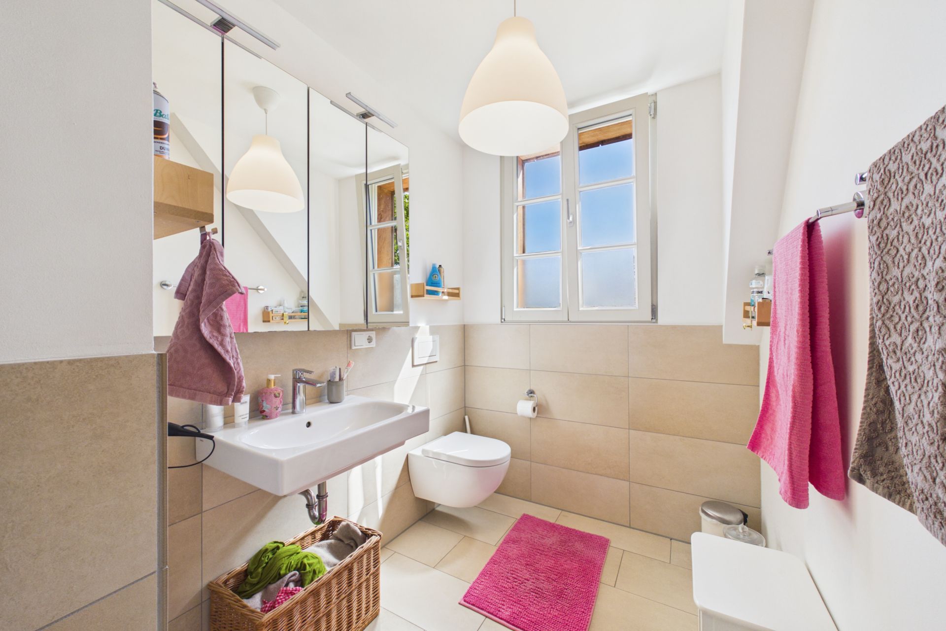 Helles Badezimmer mit Fenster, Waschbecken, WC, Spiegel und rosa Handtüchern.