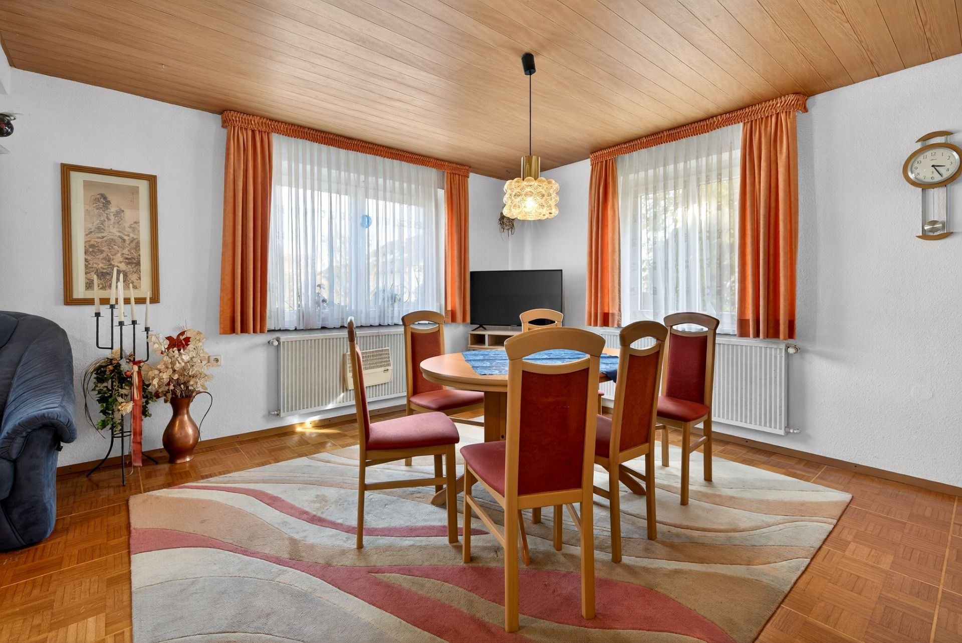 Esszimmer mit rundem Tisch, sechs Stühlen, Teppich, Fenster mit orangefarbenen Vorhängen und Fernseher.