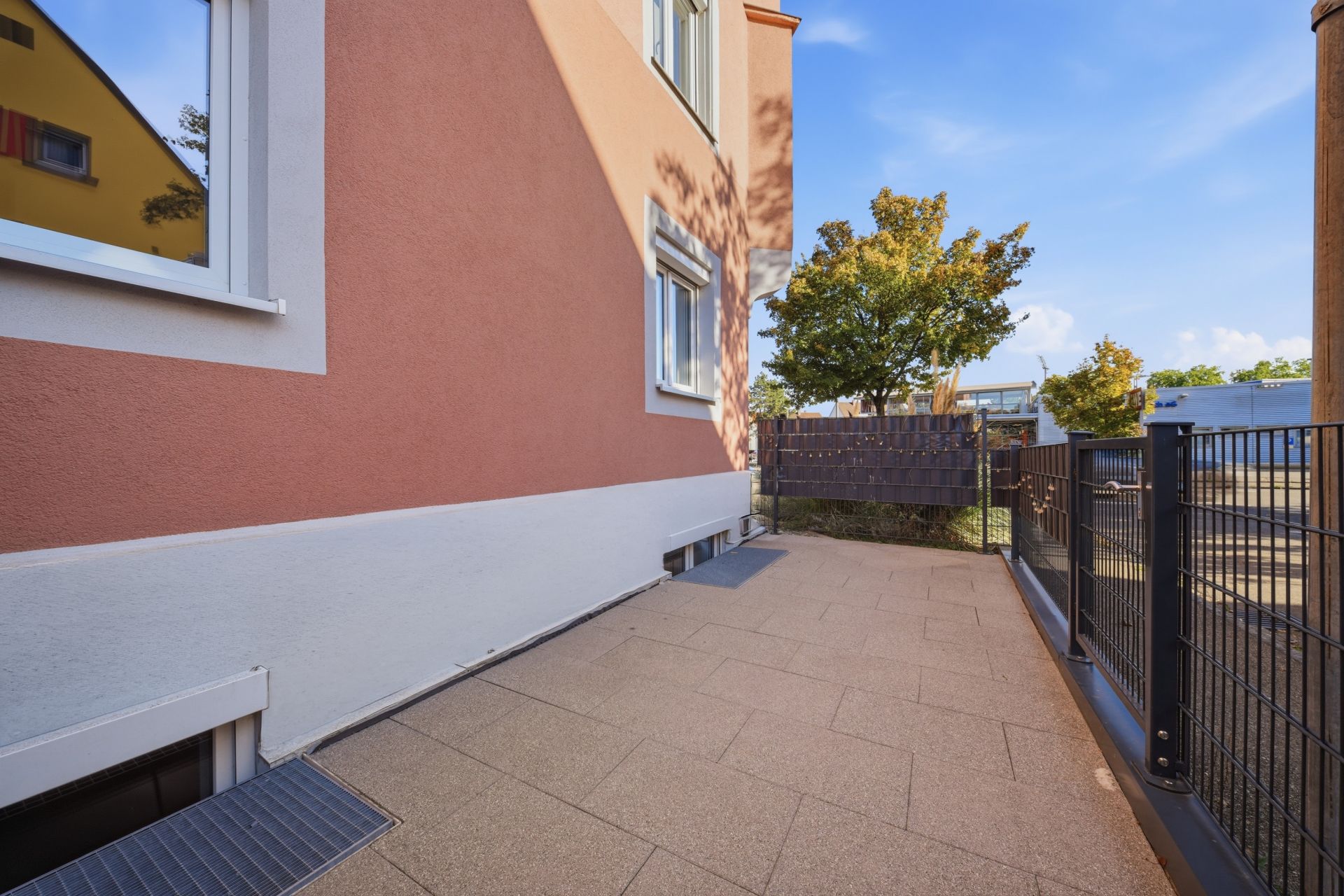 Gepflasterte Terrasse neben einem Haus mit Zaun und Bäumen im Hintergrund.