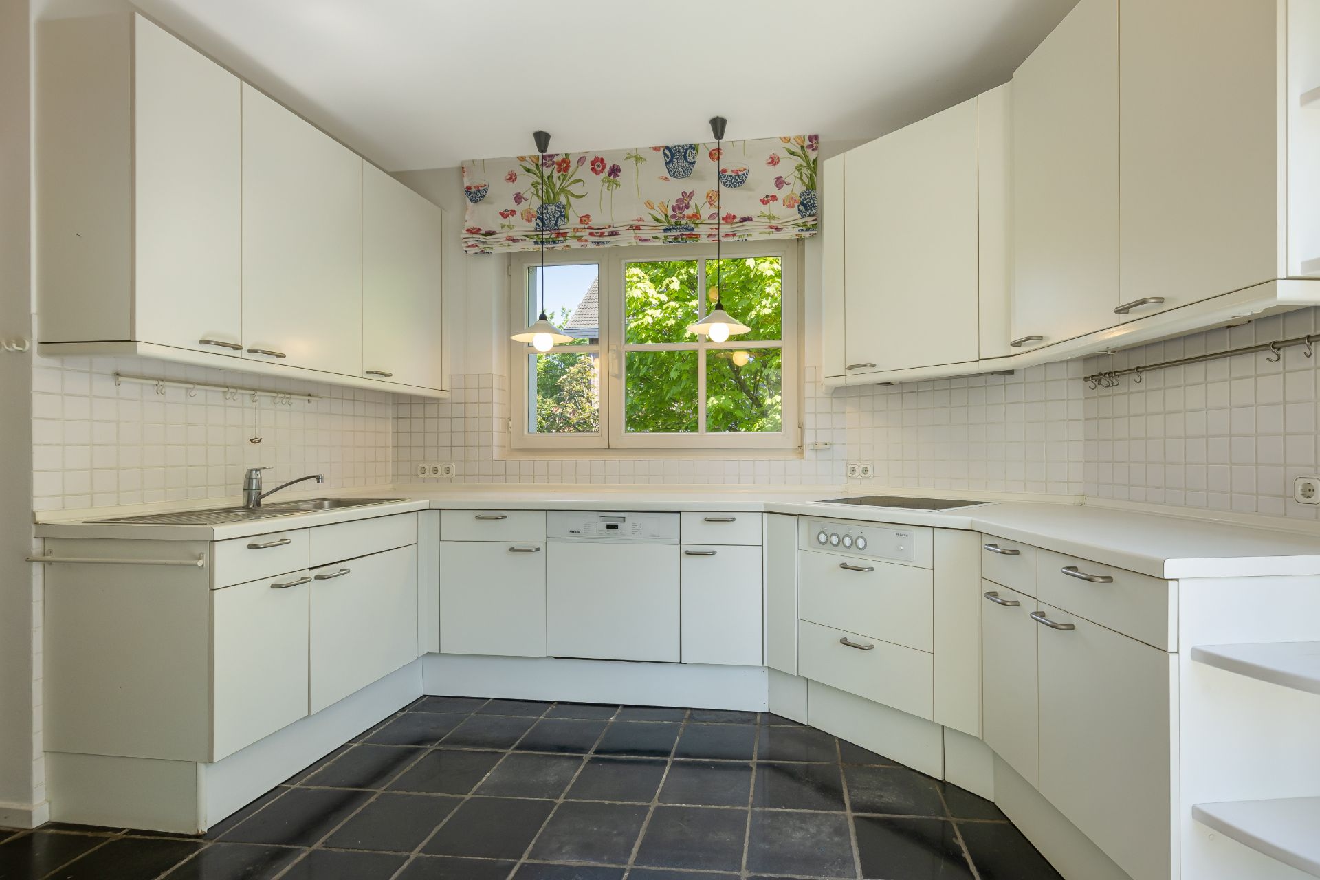Helle Einbauküche mit weißen Schränken, Fenster, dunklem Boden und zwei Hängelampen.