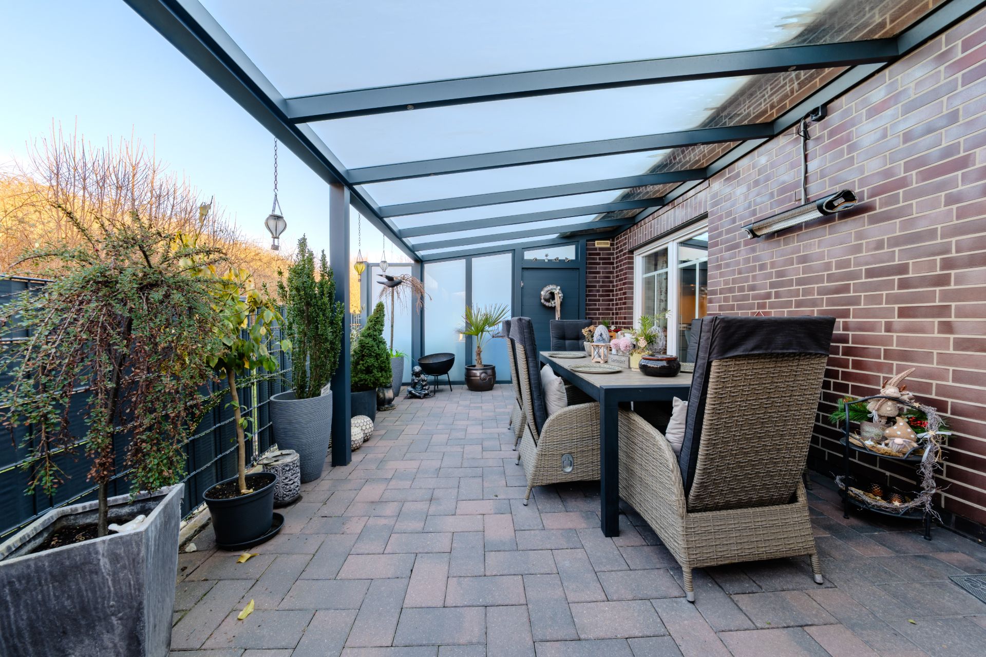 Überdachte Terrasse mit Esstisch, Stühlen und vielen Pflanzkübeln neben einer Backsteinwand.