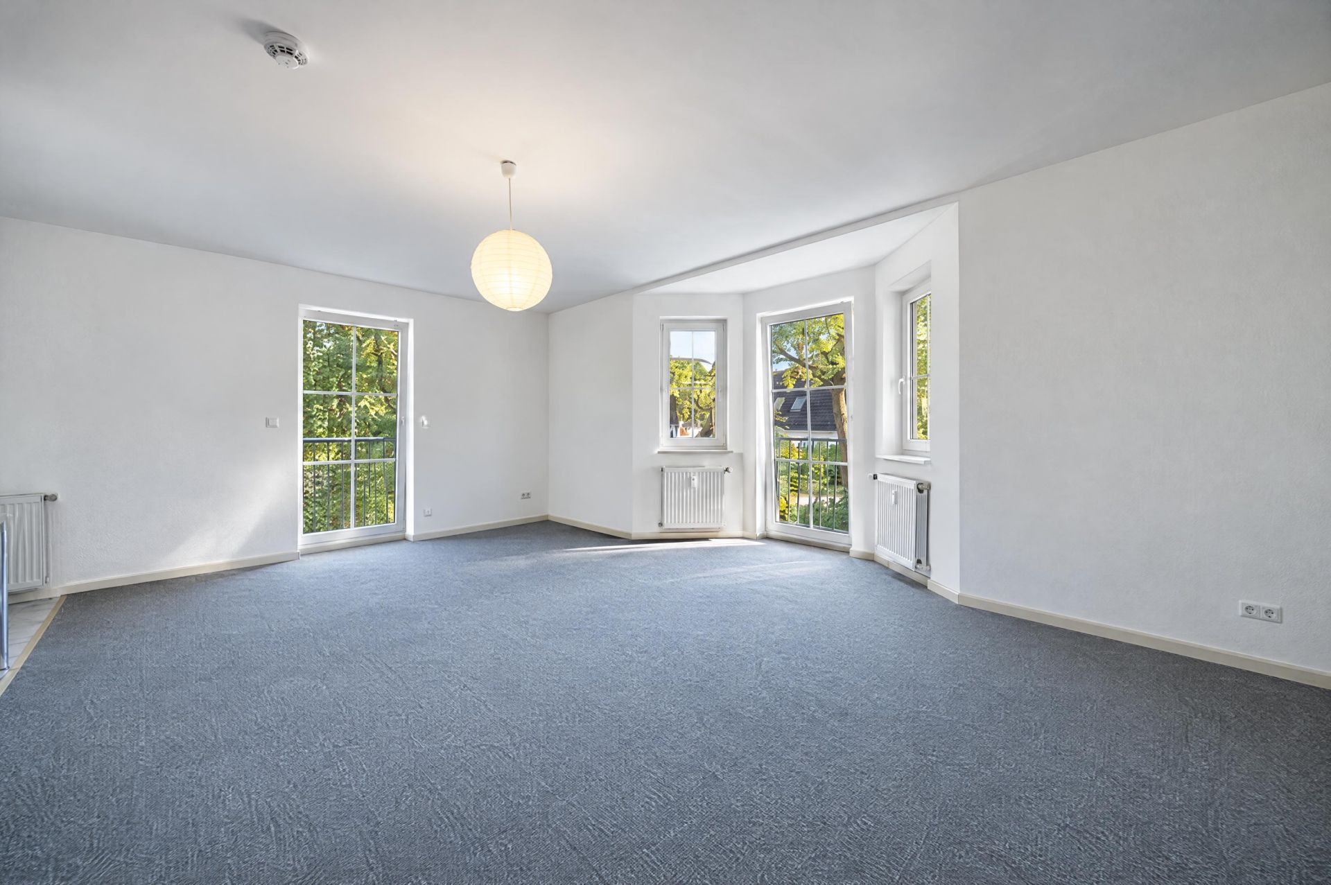 Helles, leeres Wohnzimmer mit grauem Teppich, weißen Wänden und großen Fenstern.