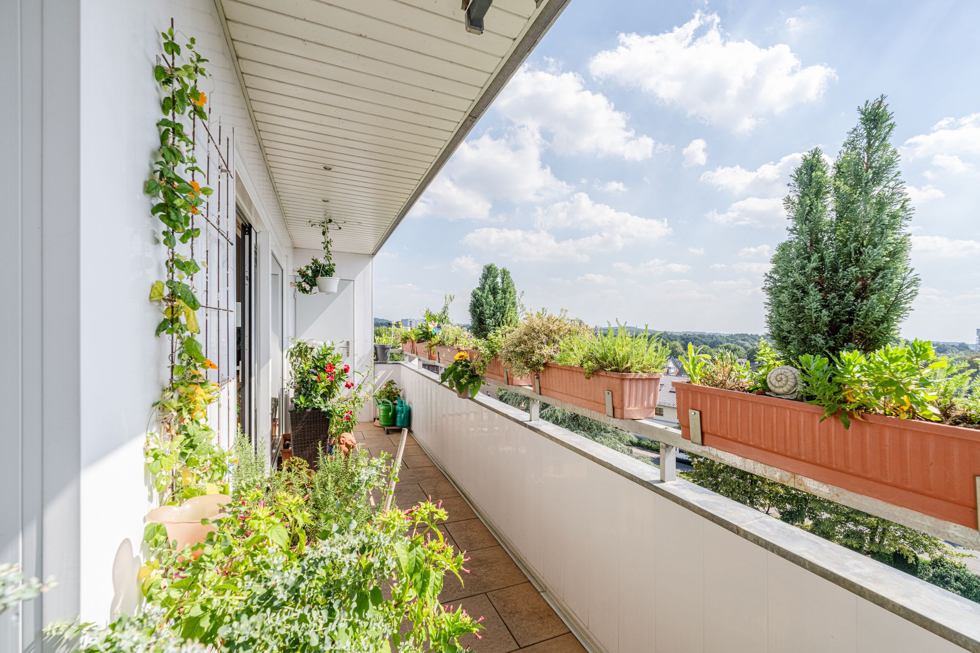 Langer Balkon mit vielen grünen Pflanzen und Blumenkästen, Blick auf Landschaft im Hintergrund.