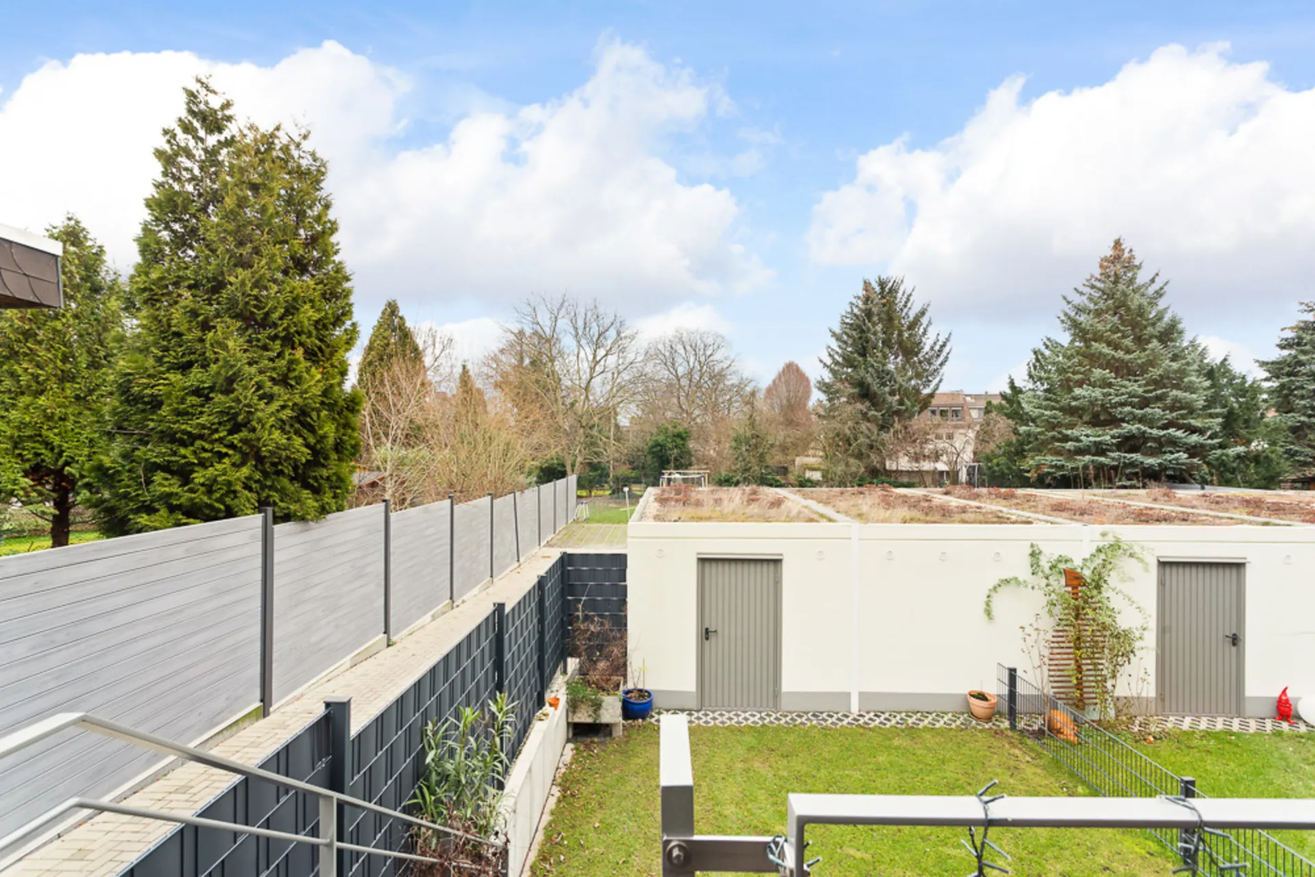 Blick auf Garten, Rasenfläche, weiße Garage mit Türen und hohe Bäume im Hintergrund.
