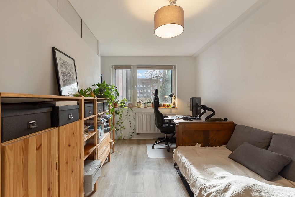 Helles Zimmer mit Schreibtisch, Sofa, Regal und Pflanzen am Fenster.