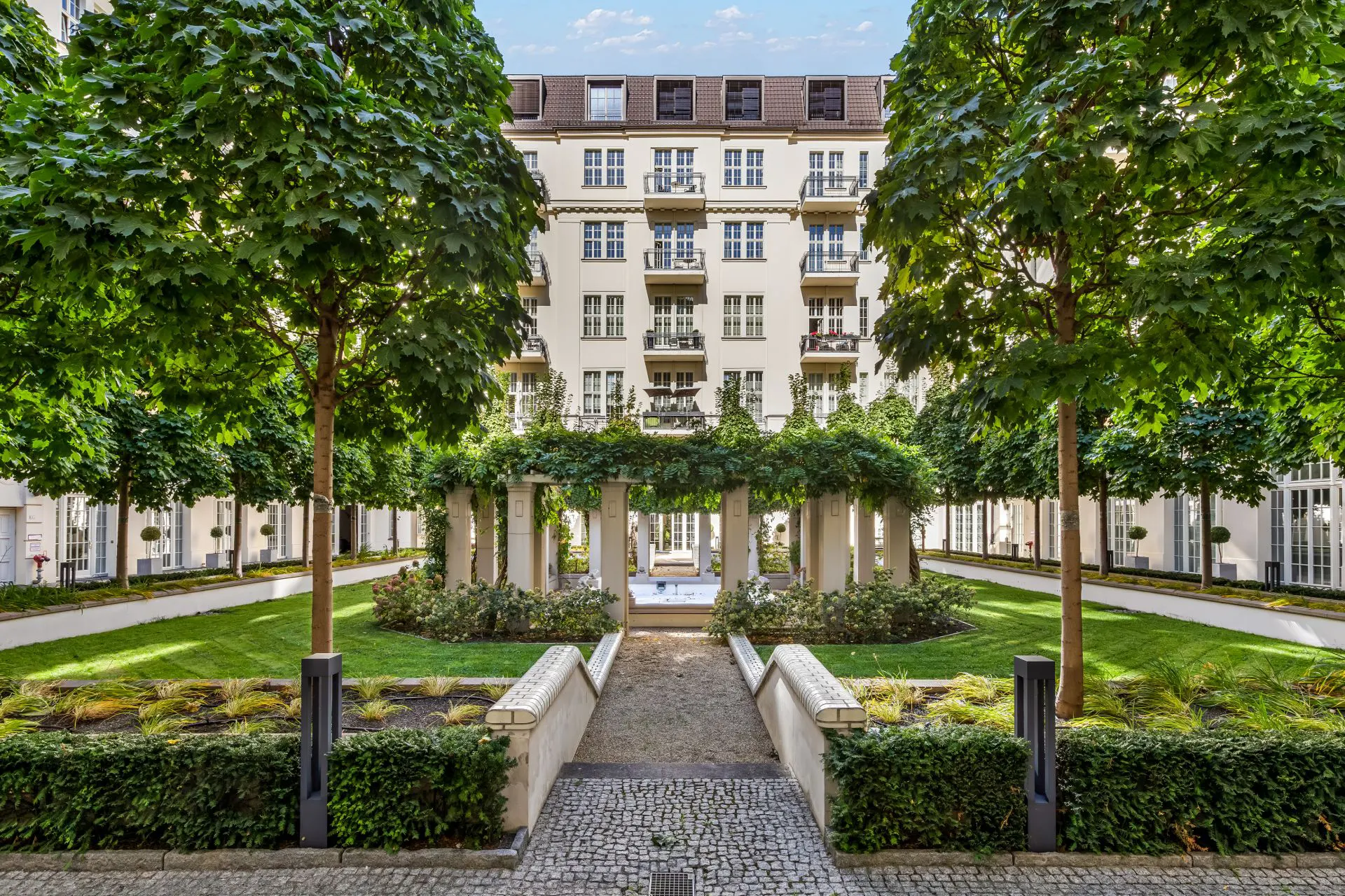Grüner Innenhof mit Bäumen, Rasenflächen, Wegen und einem Wohngebäude im Hintergrund.