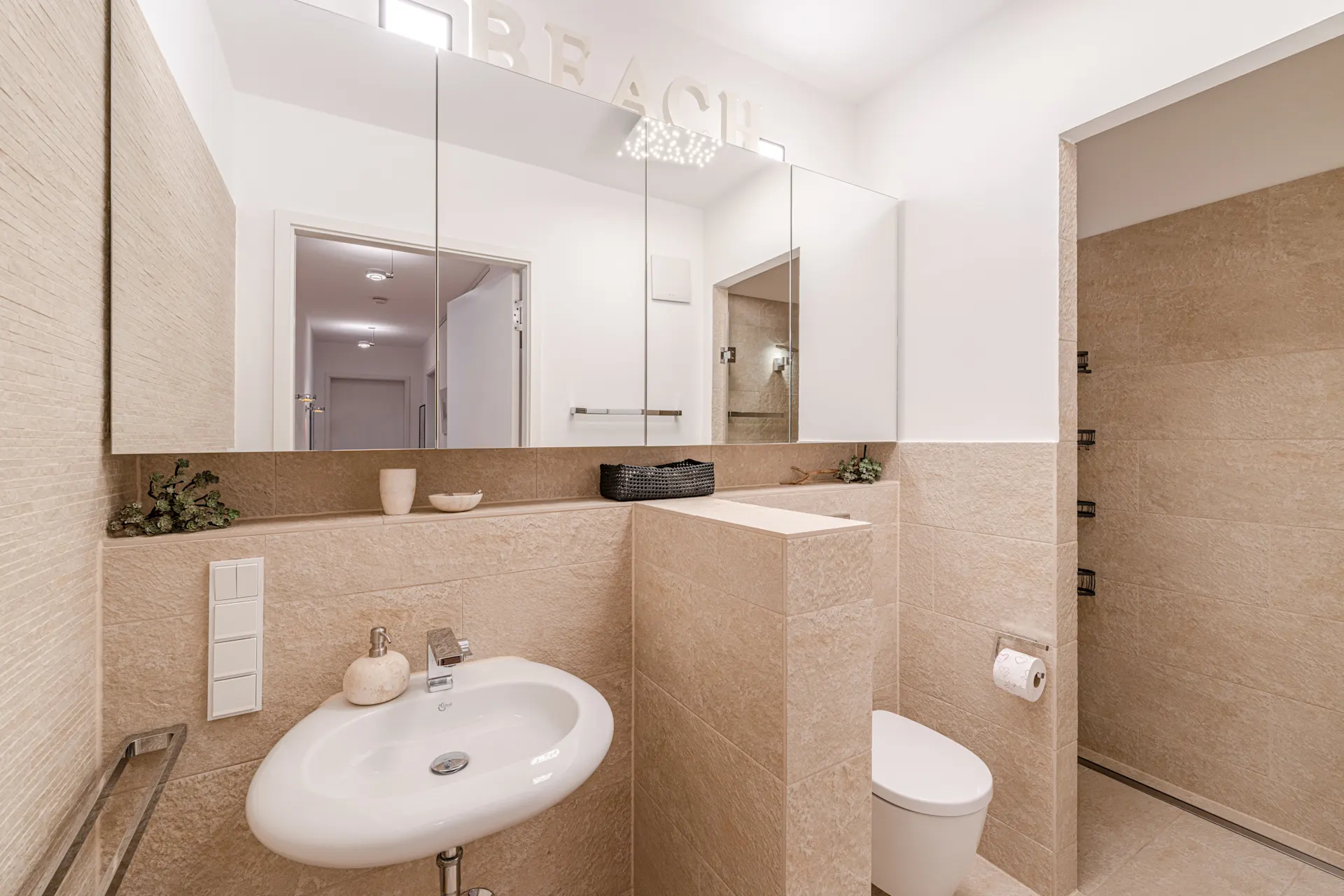 Modernes Badezimmer mit beigem Fliesen, Waschbecken, Toilette, Spiegel und offener Dusche.