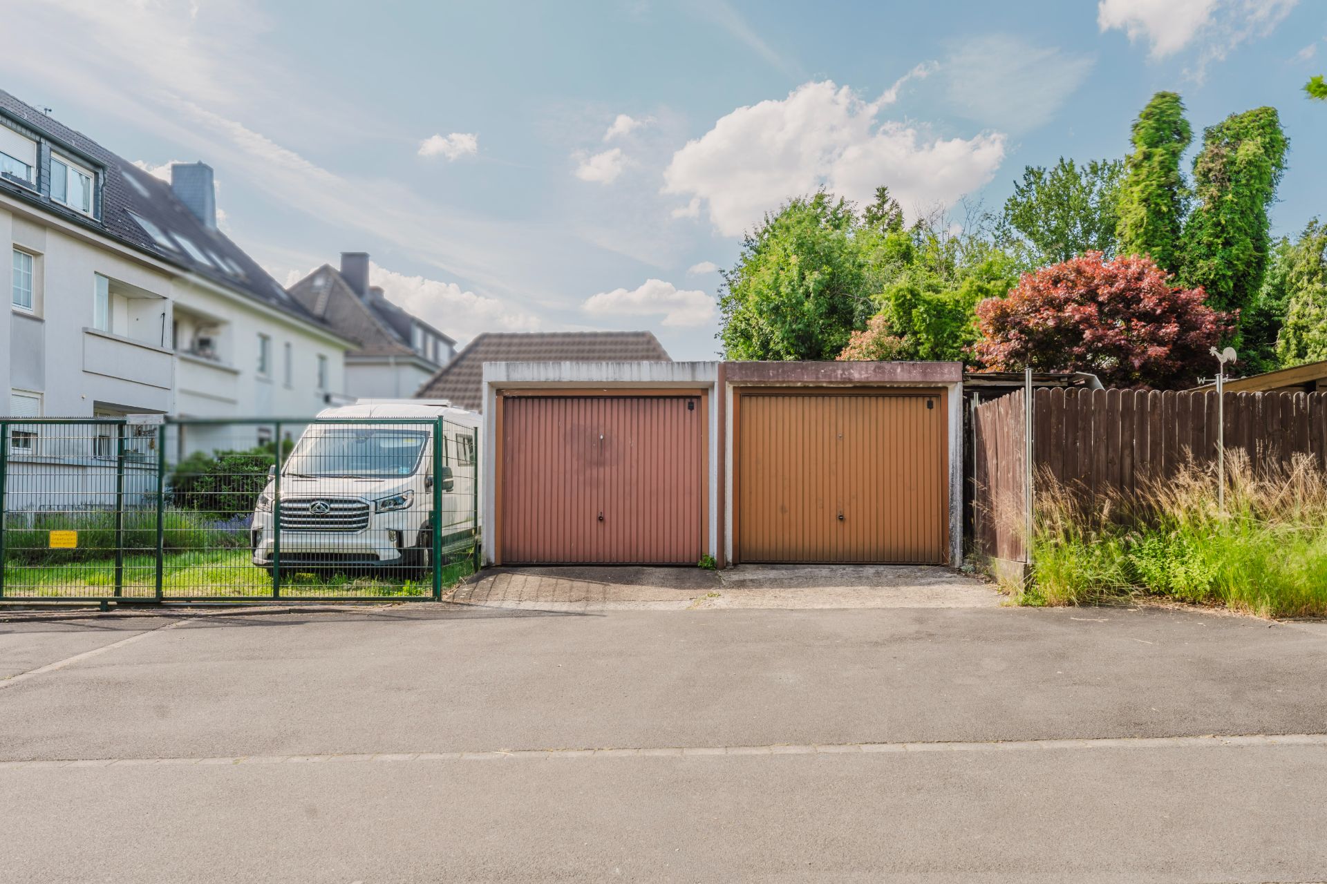 Zwei Garagen mit braunen Toren und ein weißer Transporter neben einem Zaun vor einem Wohnhaus.
