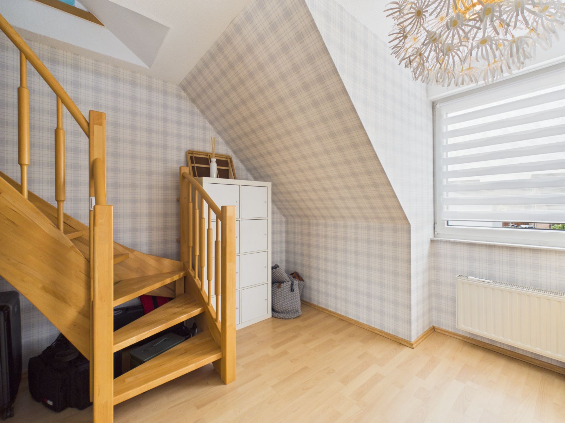 Helles Zimmer mit Holztreppe, Kommode, Fenster mit Jalousie und schräger Decke.