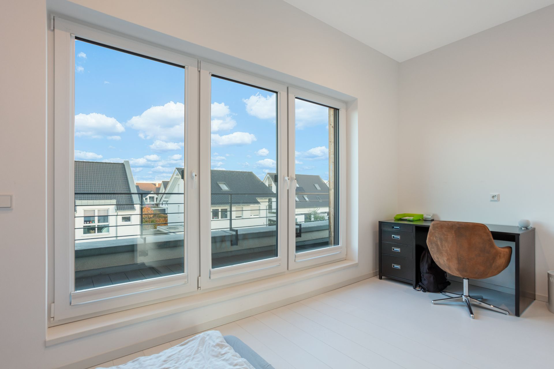 Helles Zimmer mit großem Fenster, Schreibtisch und Stuhl, Blick auf Nachbarhäuser.