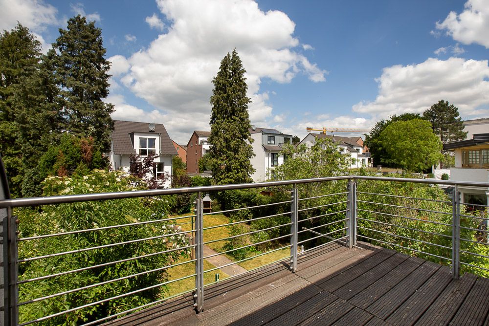 Holzbalkon mit Metallgeländer, Blick auf Gärten und Wohnhäuser bei sonnigem Wetter.