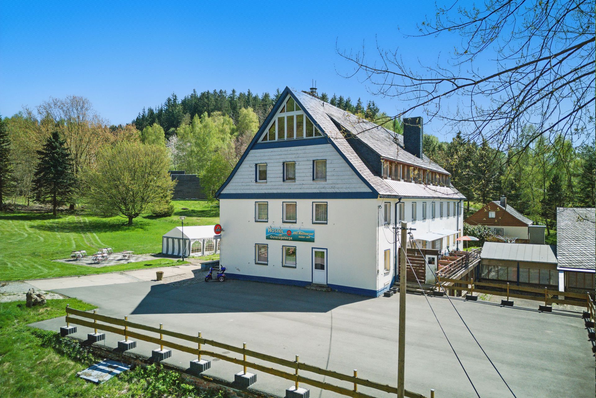 Großes Wohnhaus mit weißer Fassade, davor asphaltierter Hof, umgeben von grüner Wiese und Bäumen.