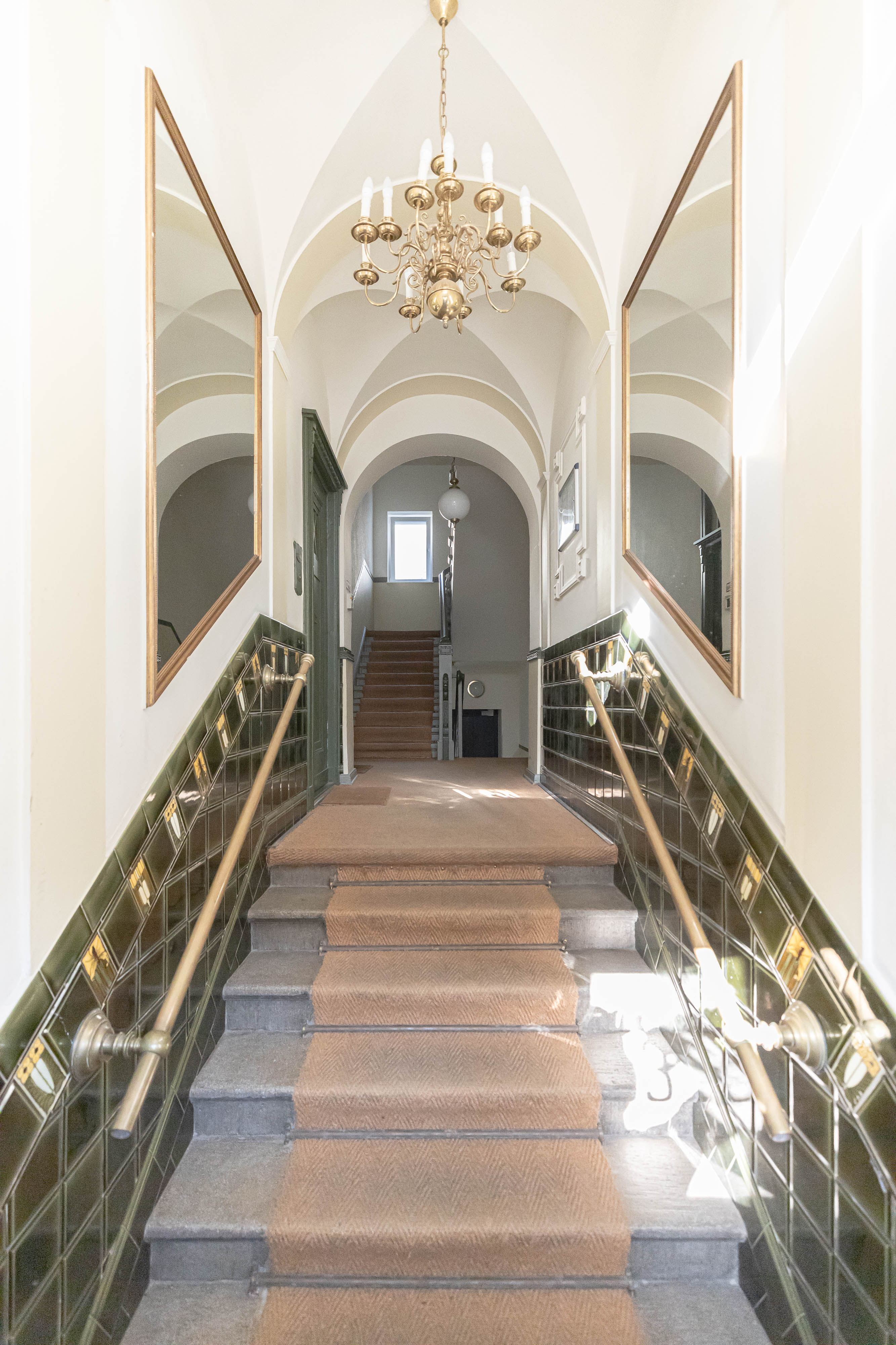 Heller Hausflur mit Teppichtreppe, Kronleuchter und zwei großen Spiegeln an den Wänden.