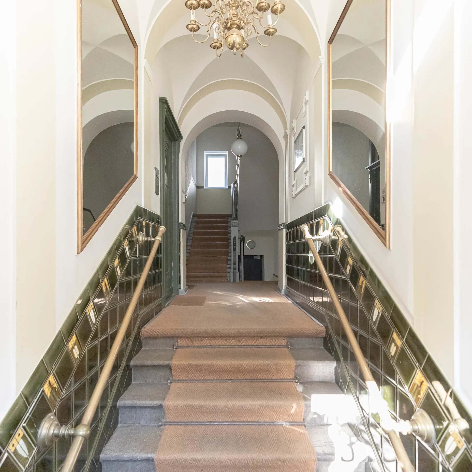 Heller Hausflur mit Teppichtreppe, Kronleuchter und zwei großen Spiegeln an den Wänden.