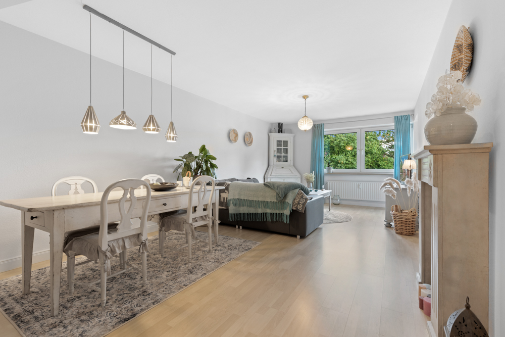 Helles Wohn- und Esszimmer mit Holzboden, Esstisch, Sofa, großen Fenstern und grünen Vorhängen.