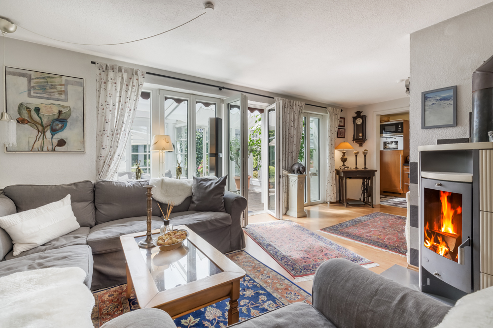 Helles Wohnzimmer mit grauem Sofa, Kamin, Glastüren zum Garten und Teppichen auf Holzboden.