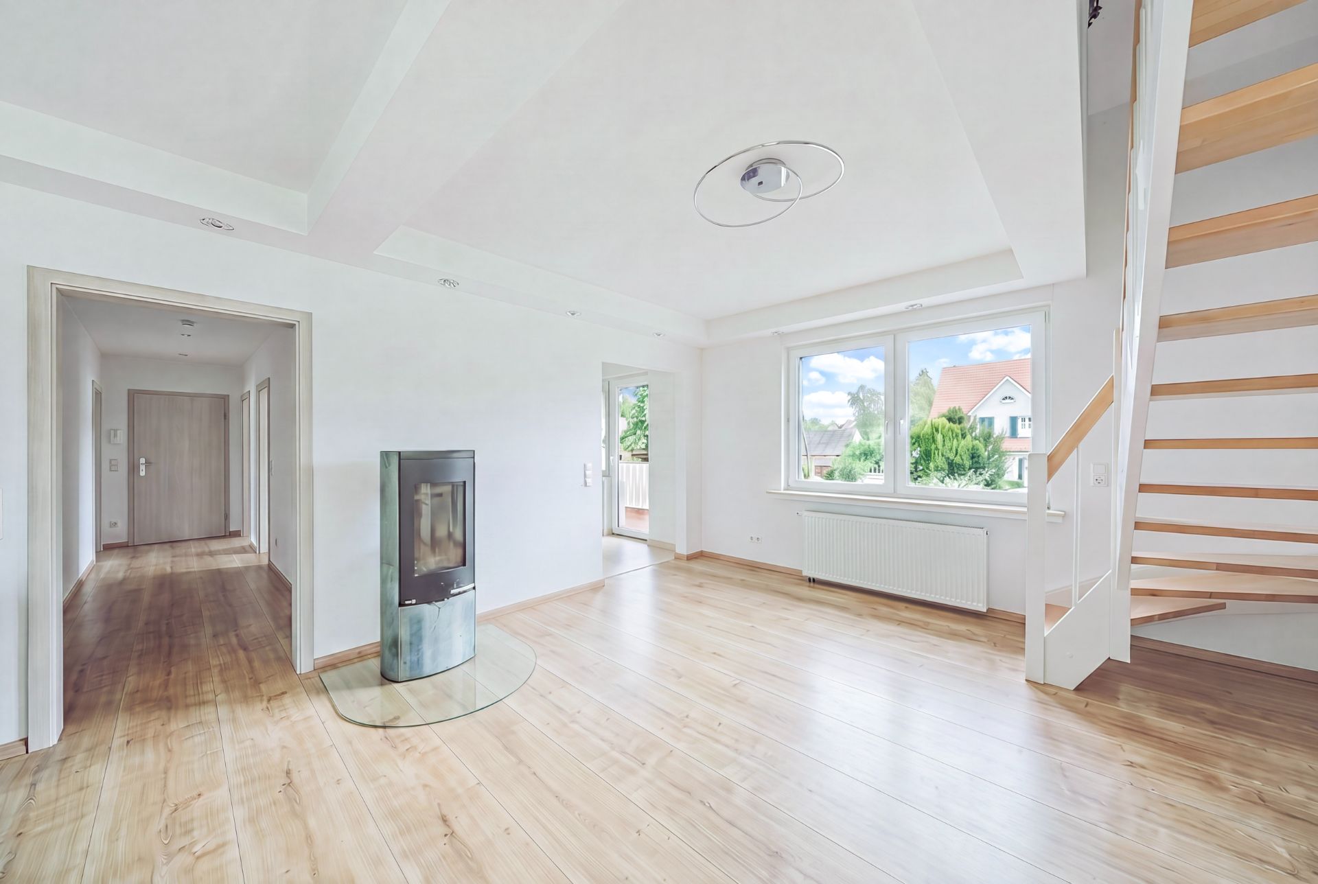 Helles Wohnzimmer mit Kaminofen, großem Fenster, Holztreppe und Flur.