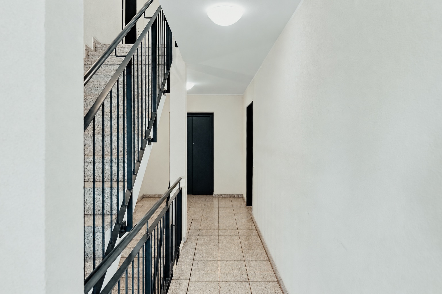 Heller Flur mit Fliesenboden, schwarzen Türen und einer Treppe mit Metallgeländer.