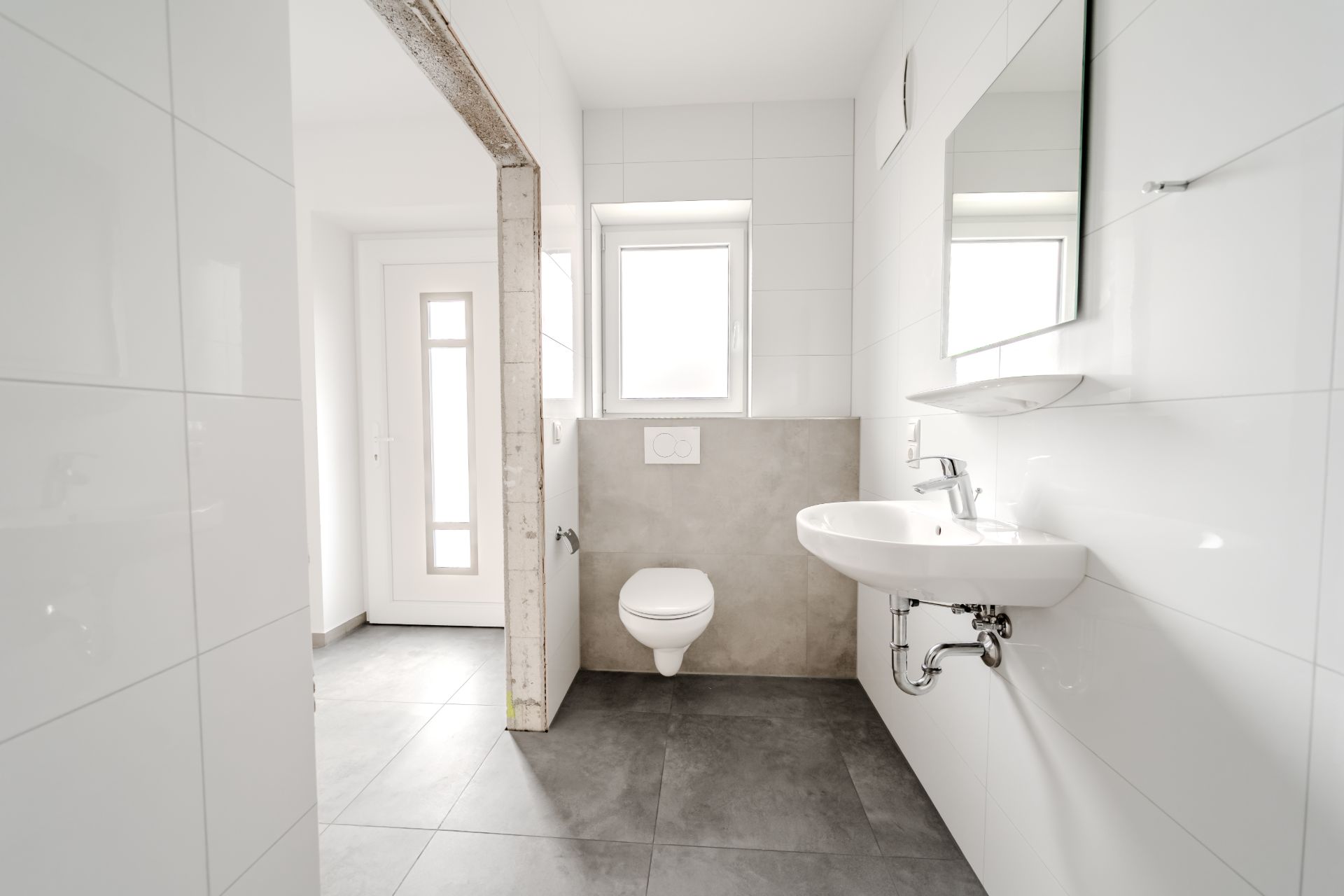 Helles Badezimmer mit Wandtoilette, Waschbecken, Spiegel und Fenster.