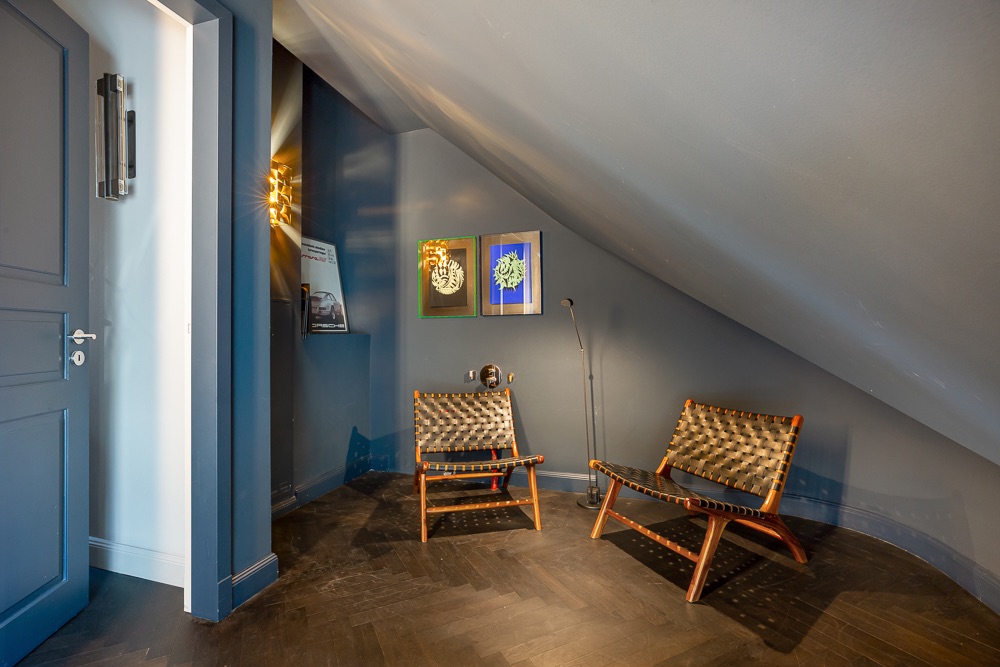 Zwei geflochtene Stühle stehen unter einer schrägen Decke, daneben hängen bunte Bilder an der blauen Wand.
