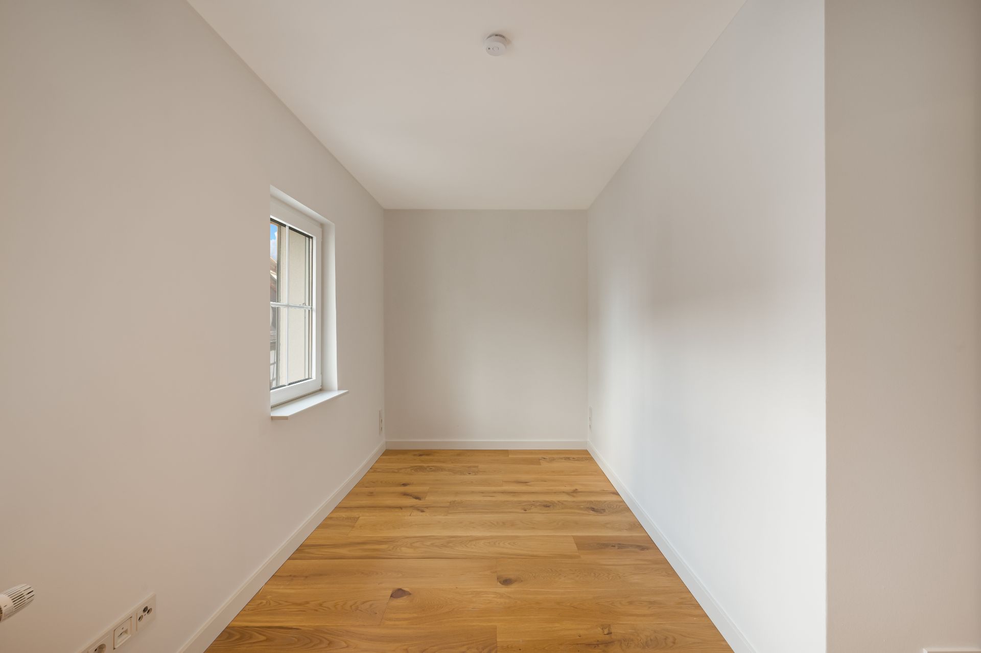 Leerer, schmaler Raum mit Holzboden und einem Fenster an der linken Wand.