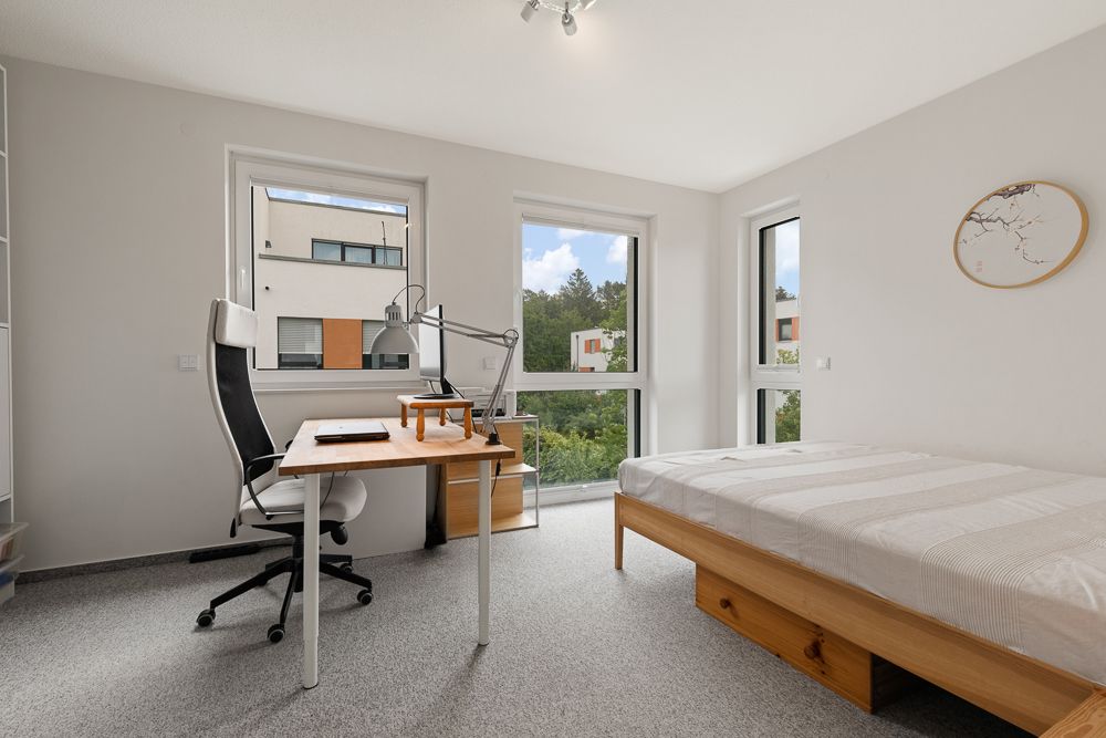 Helles Schlafzimmer mit Schreibtisch, Stuhl und Einzelbett, große Fenster mit Blick ins Grüne.