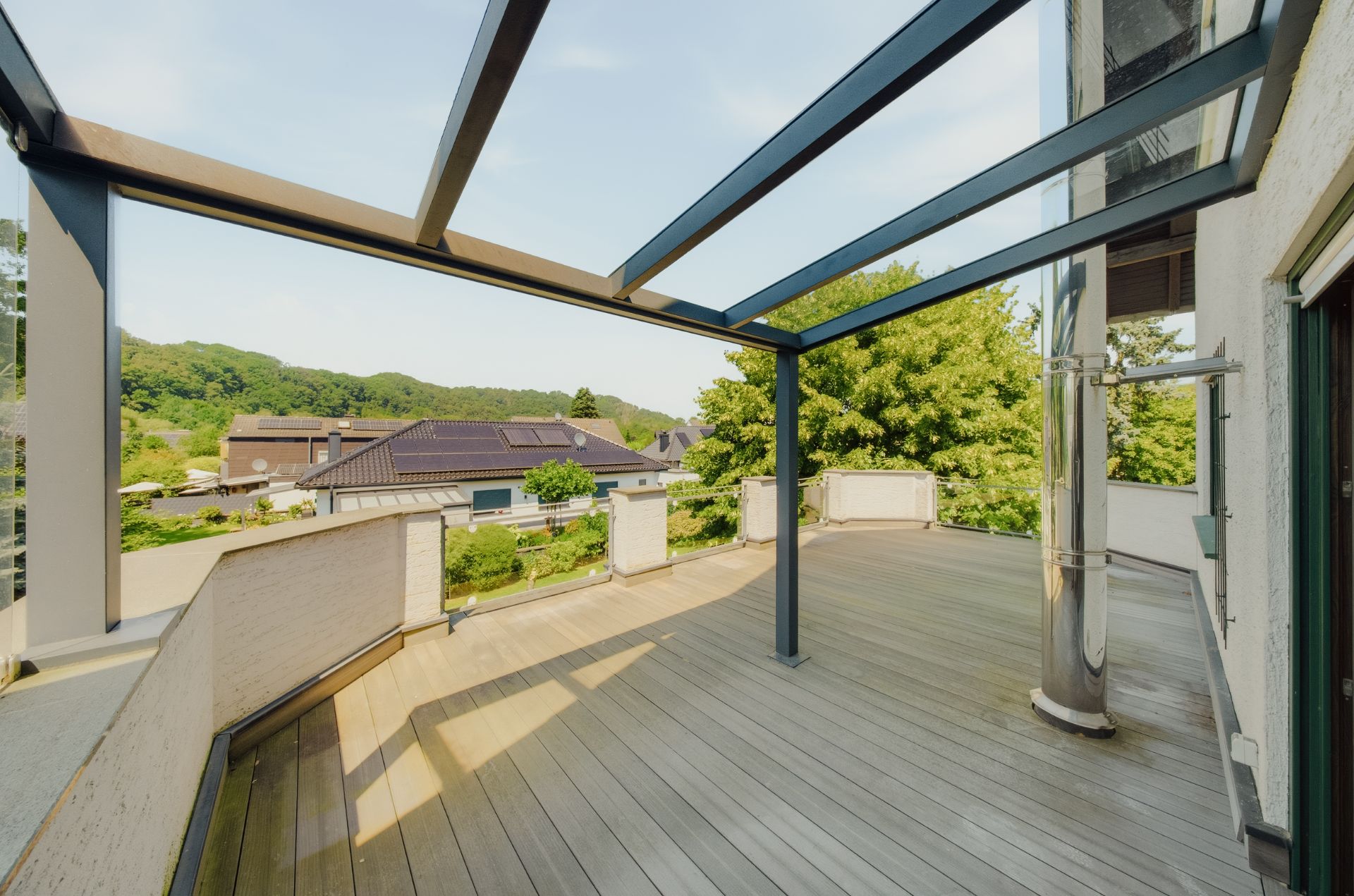 Große Terrasse mit Holzboden, Glasdachrahmen und Blick auf grüne Bäume und Nachbarhäuser.