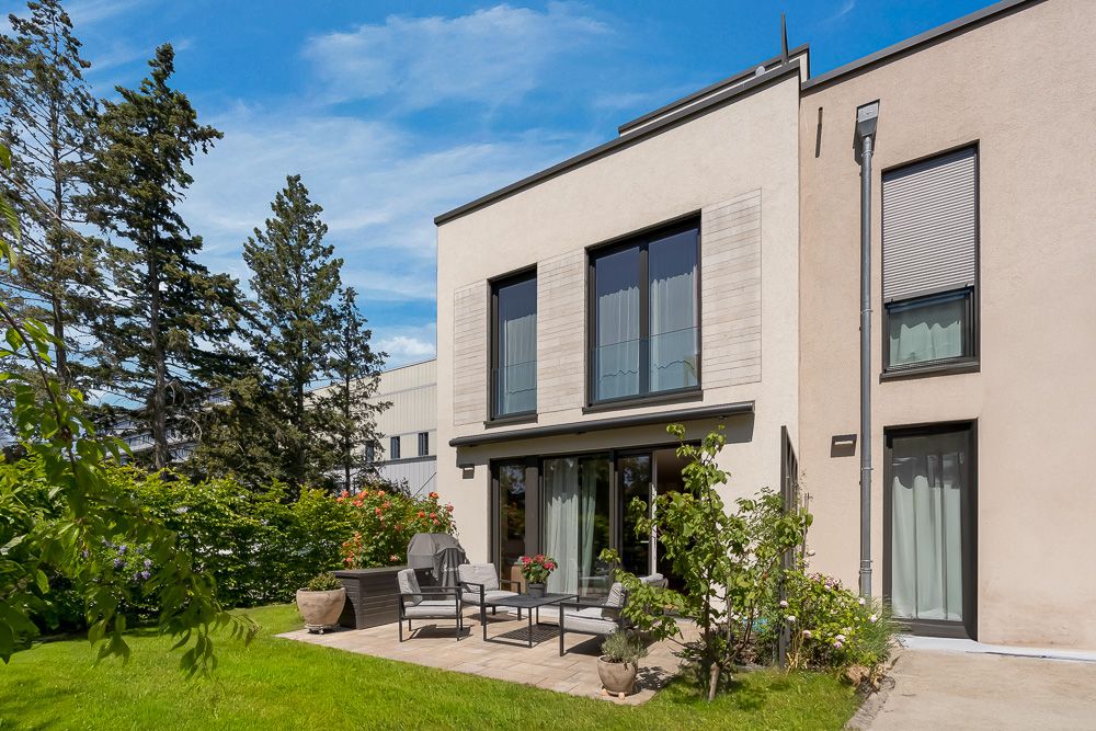 Moderne Wohnhausfassade mit Terrasse, Gartenmöbeln und gepflegtem Garten bei sonnigem Wetter.