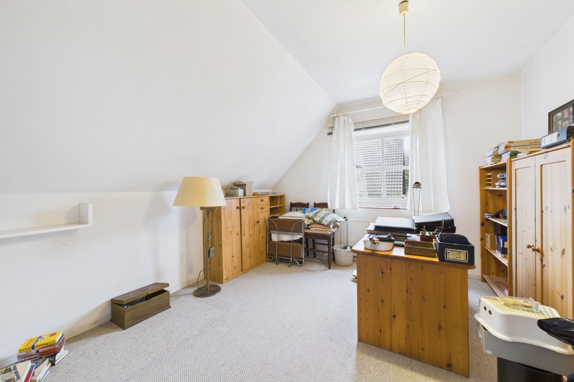 Helles Zimmer mit Holzschreibtisch, Regalen, Stuhl, Stehlampe und Fenster mit weißen Vorhängen.