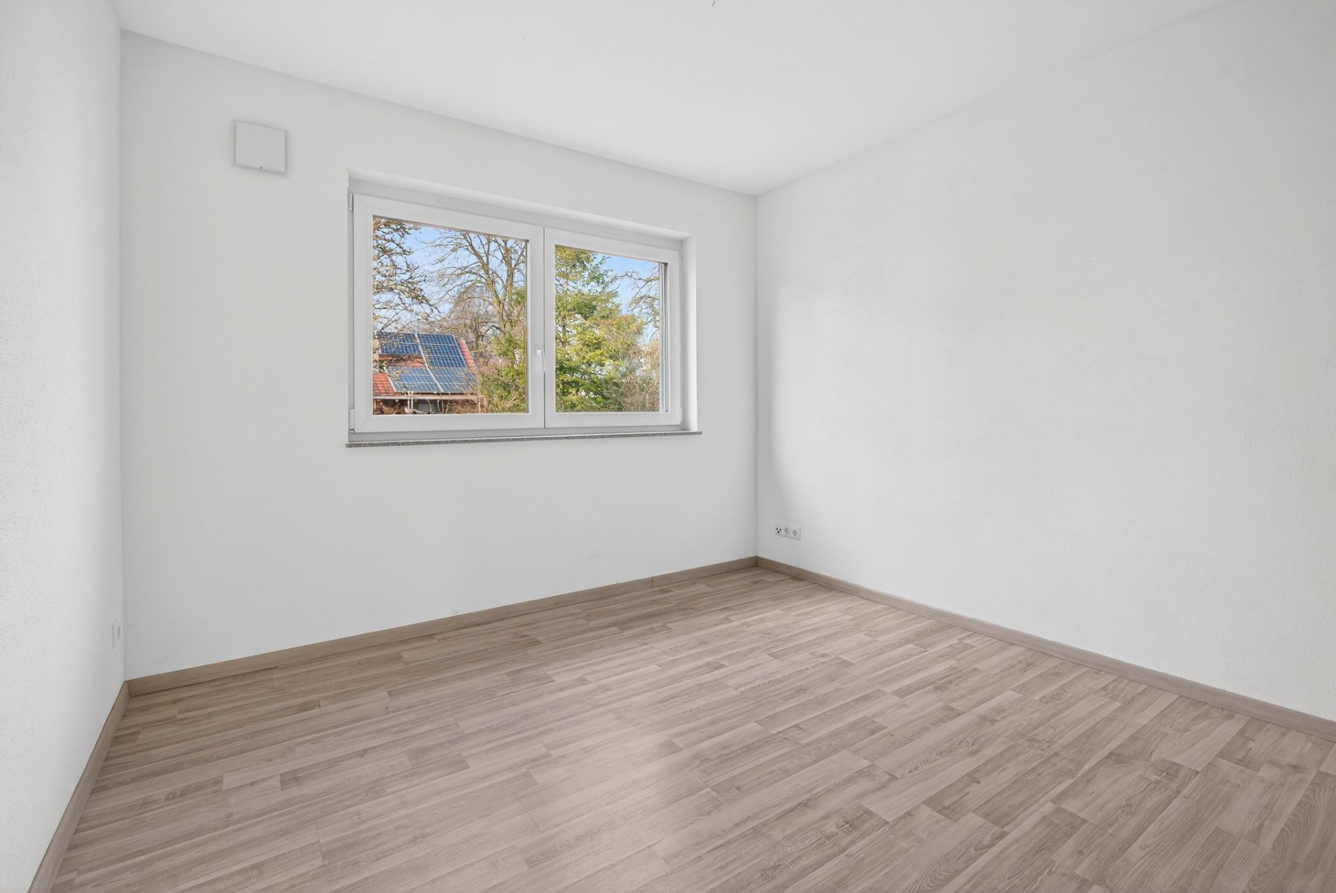 Leeres Zimmer mit hellem Holzfußboden und großem Fenster, weiße Wände.