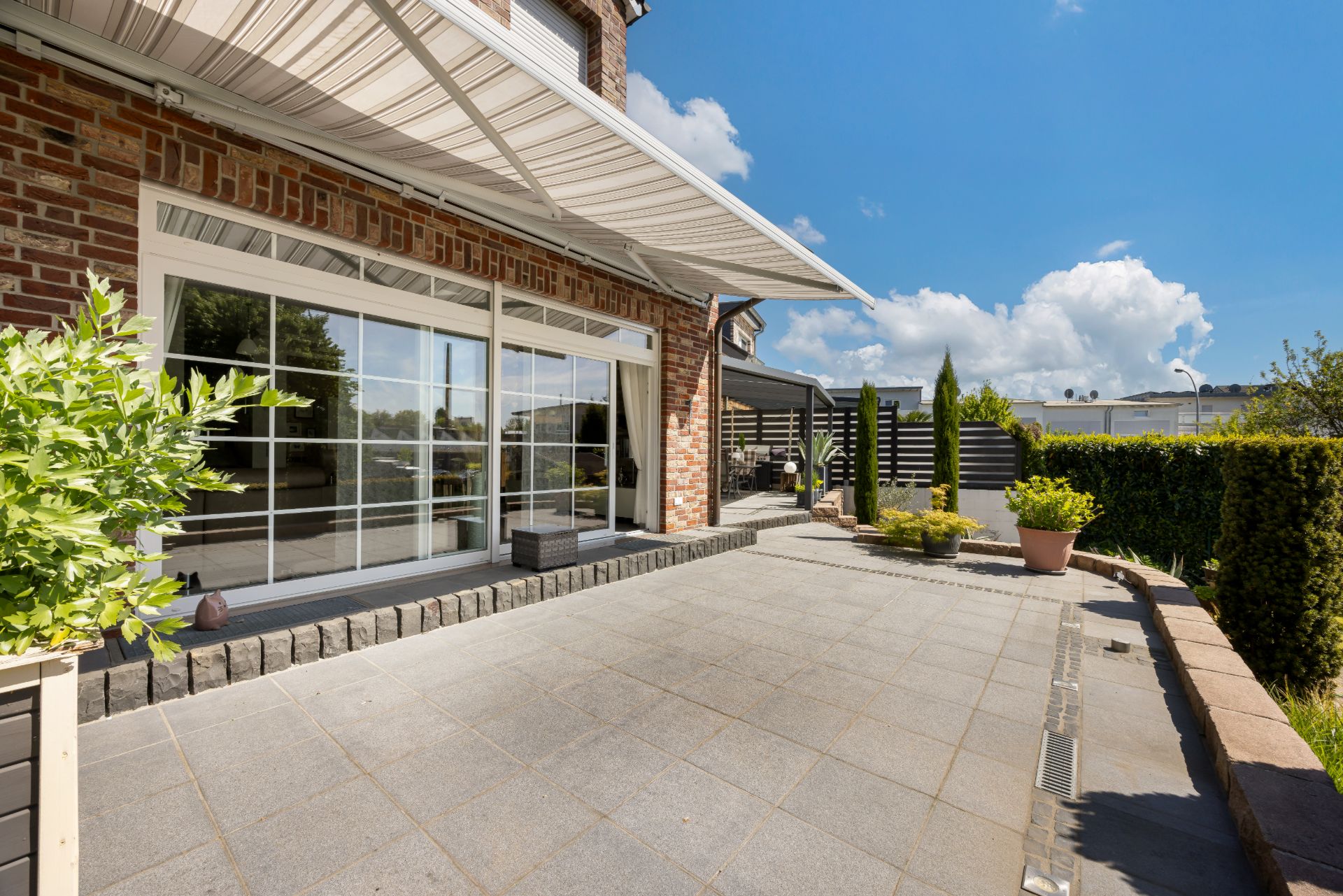 Große Terrasse mit Fliesenboden, Markise und Pflanzen vor einem Wohnhaus mit Glas-Schiebetüren.