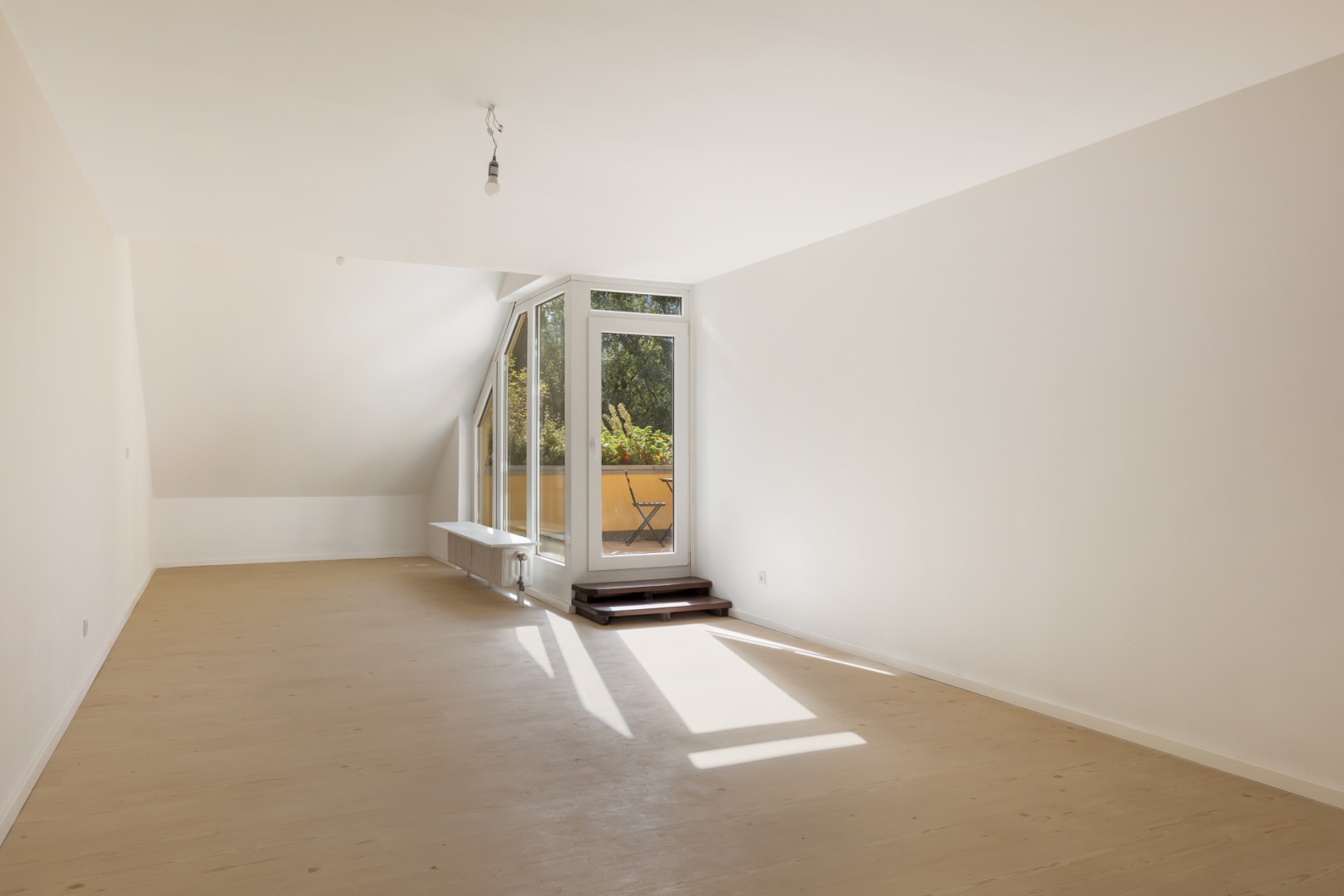 Helles, leeres Zimmer mit großen Fenstern und Tür zu einem kleinen Balkon mit Blick ins Grüne.