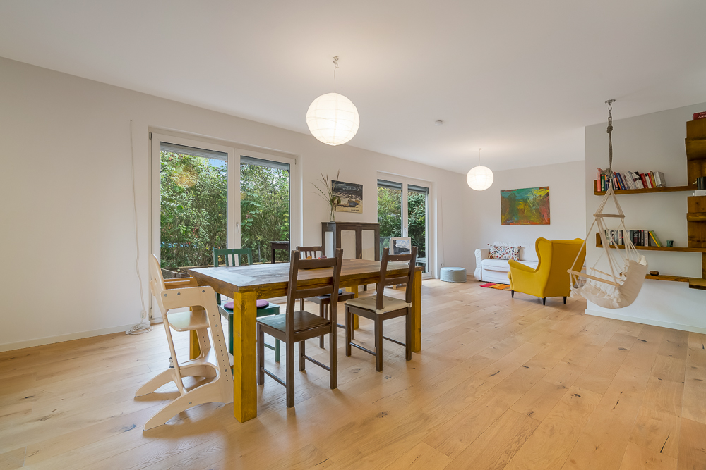 Helles Wohnzimmer mit Holzboden, Esstisch, Hängesessel, gelbem Sessel, Sofa und großen Fenstern.
