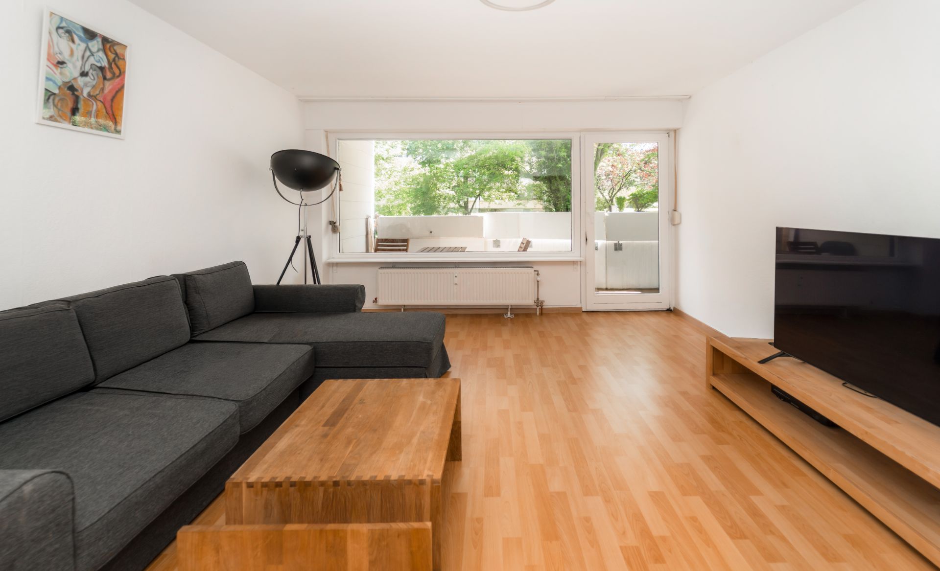 Wohnzimmer mit dunklem Ecksofa, Holztisch, Fernseher und großem Fenster mit Blick ins Grüne.