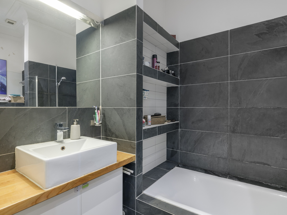 Modernes Badezimmer mit Waschbecken, Spiegel, Badewanne und dunklen Fliesen.