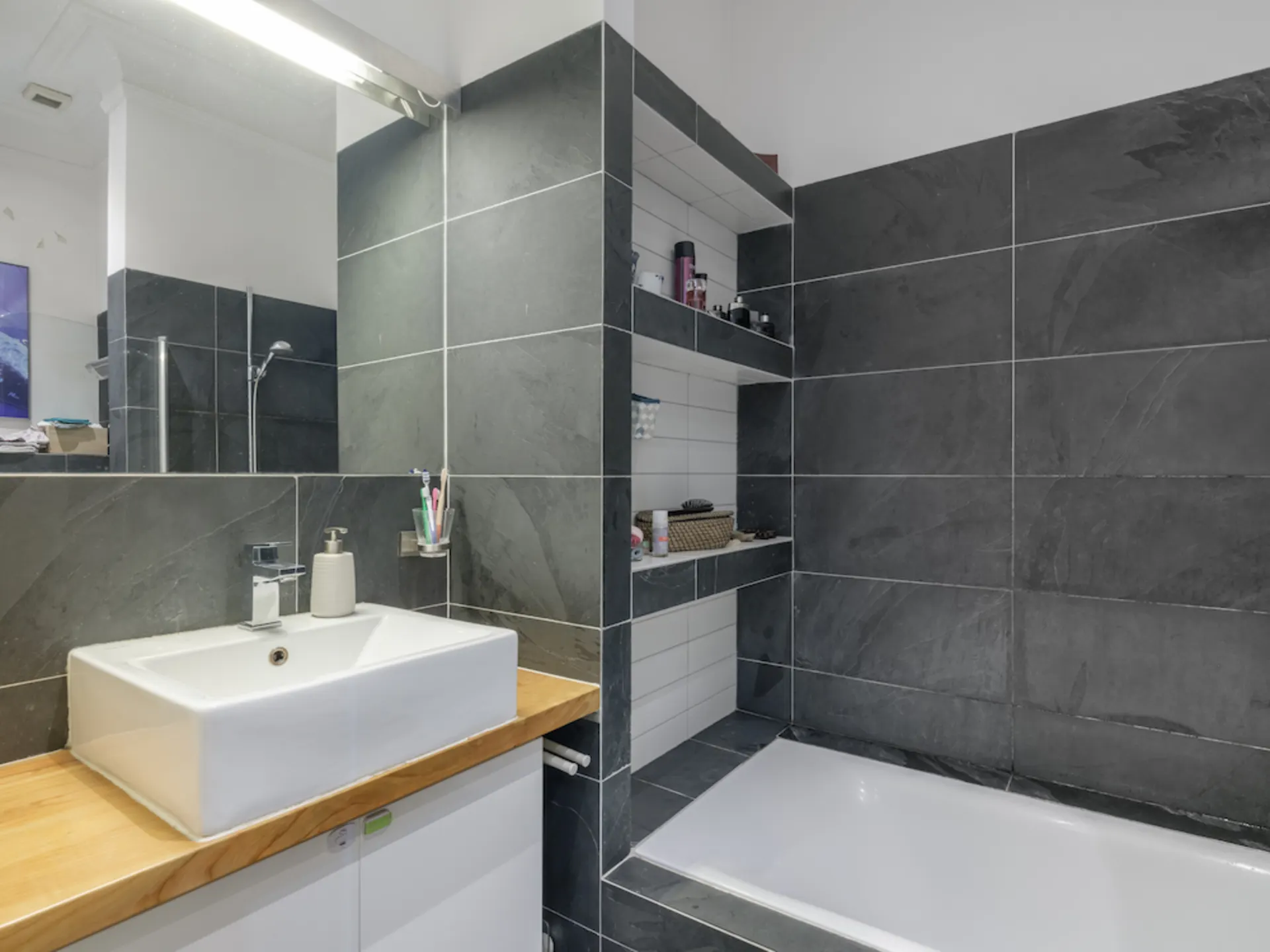 Modernes Badezimmer mit Waschbecken, Spiegel, Badewanne und dunklen Fliesen.