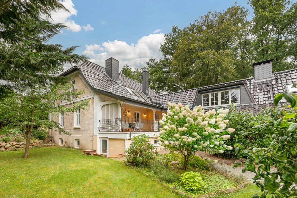 Haus mit grauem Dach, Balkon und Garten mit blühenden Sträuchern und Bäumen.