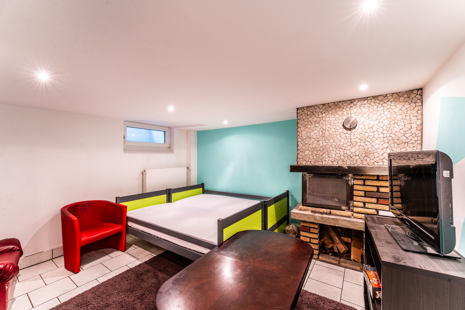 Helles Zimmer mit Bett, rotem Sessel, Kamin, Fernseher und kleinem Fenster.