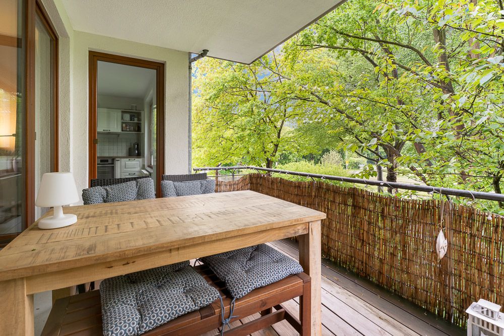 Balkon mit Holztisch, Stühlen und Blick auf grüne Bäume, Zugang zur Küche sichtbar.