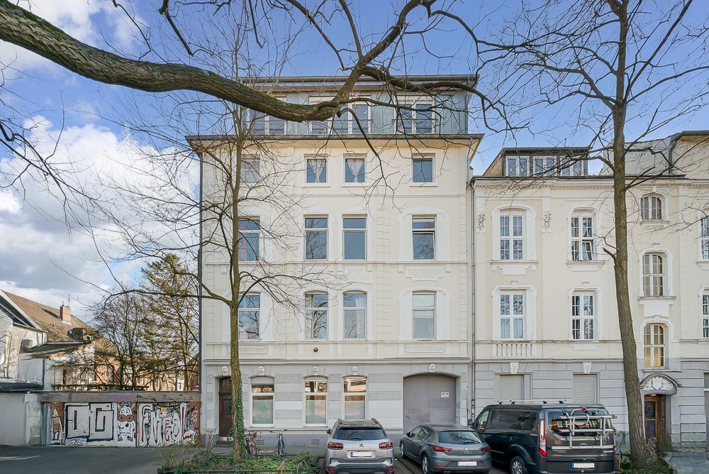 Mehrstöckiges Wohngebäude mit hellen Fassaden, geparkten Autos und Bäumen im Vordergrund.