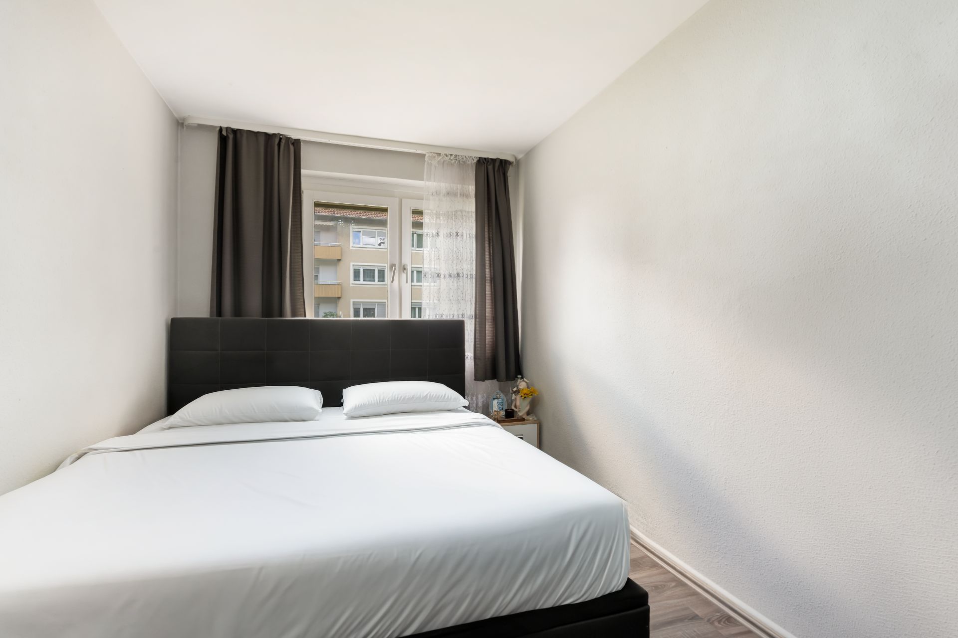 Helles Schlafzimmer mit großem Bett, Fenster, Vorhängen und Nachttisch mit Dekoration.
