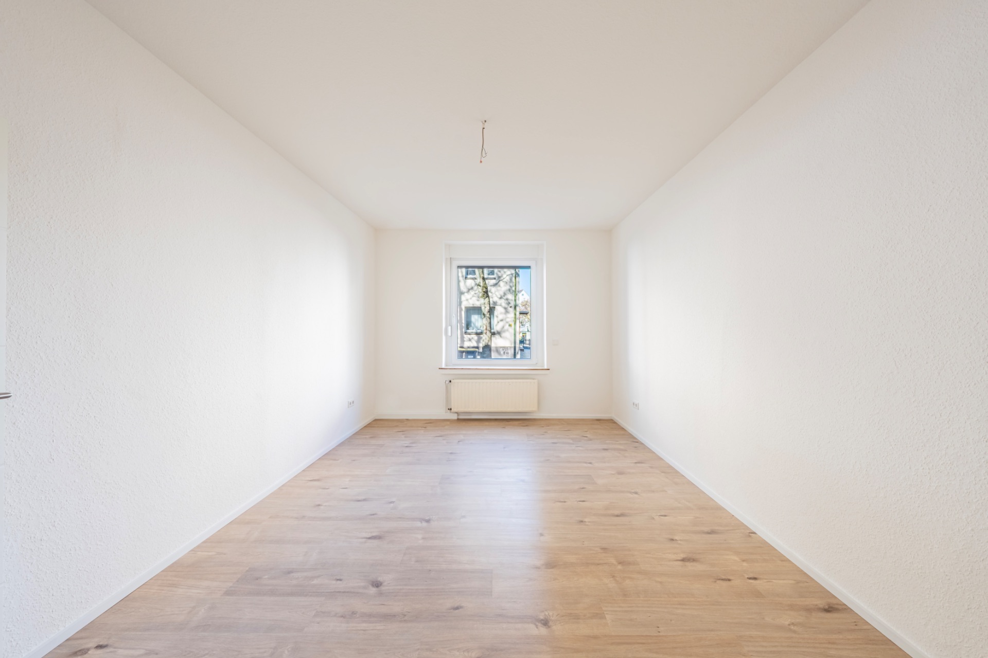 Leerer, heller Raum mit Holzfußboden, weiß gestrichenen Wänden und Fenster am Ende.
