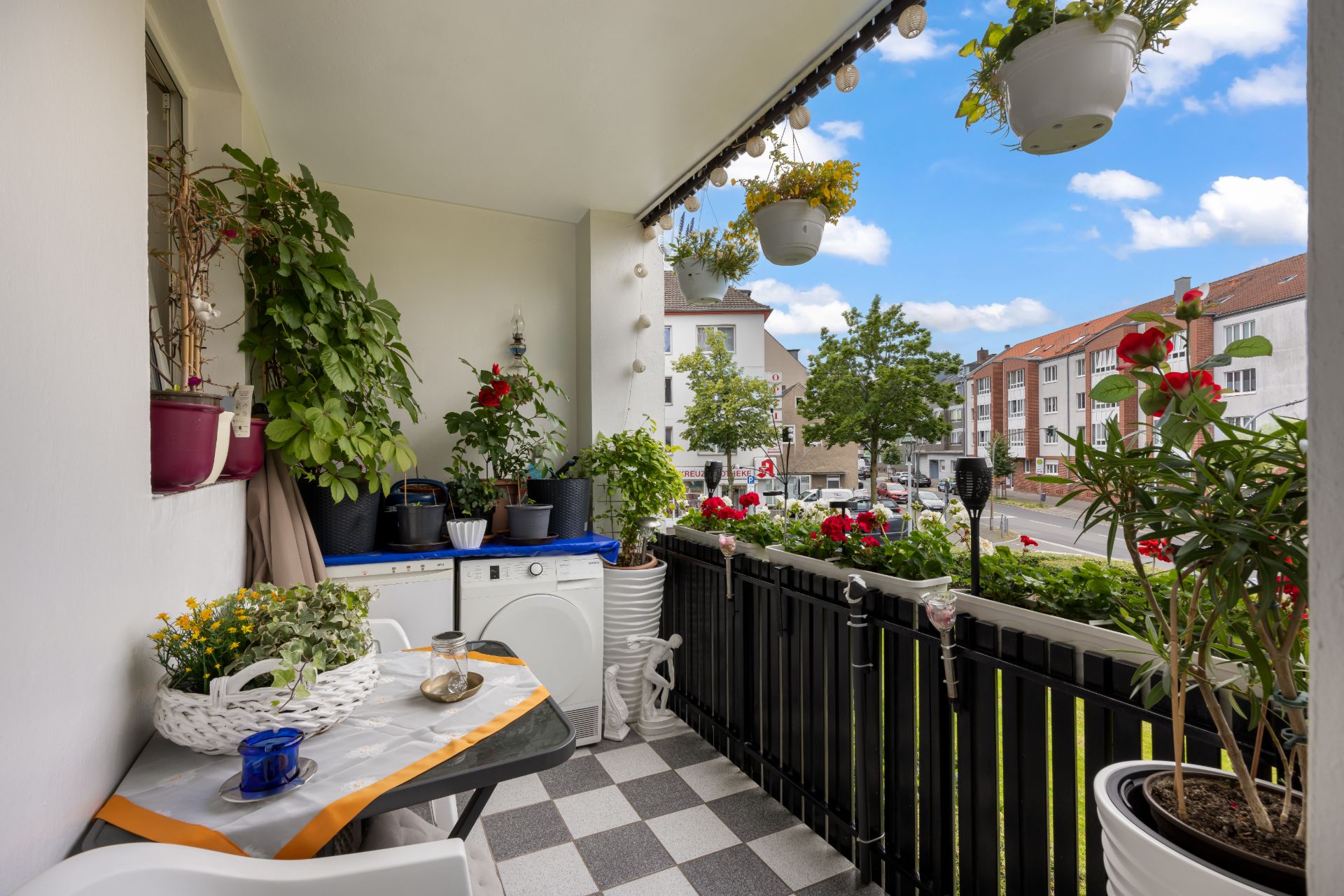 Balkon mit vielen Pflanzen, kleinem Tisch und Blick auf eine Straße mit Wohnhäusern.