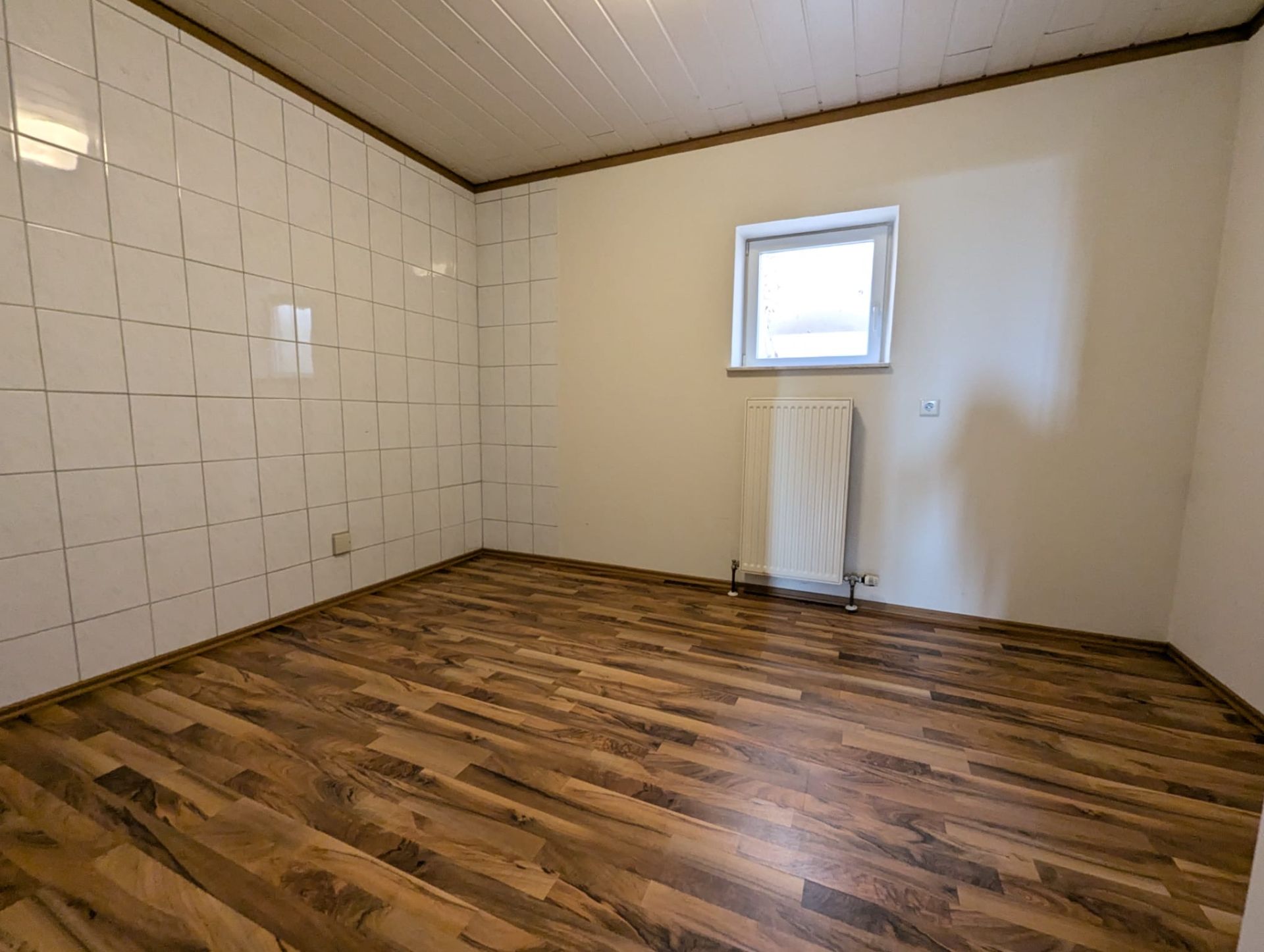 Leerer Raum mit Holzfußboden, teils gefliester Wand, kleinem Fenster und Heizkörper.