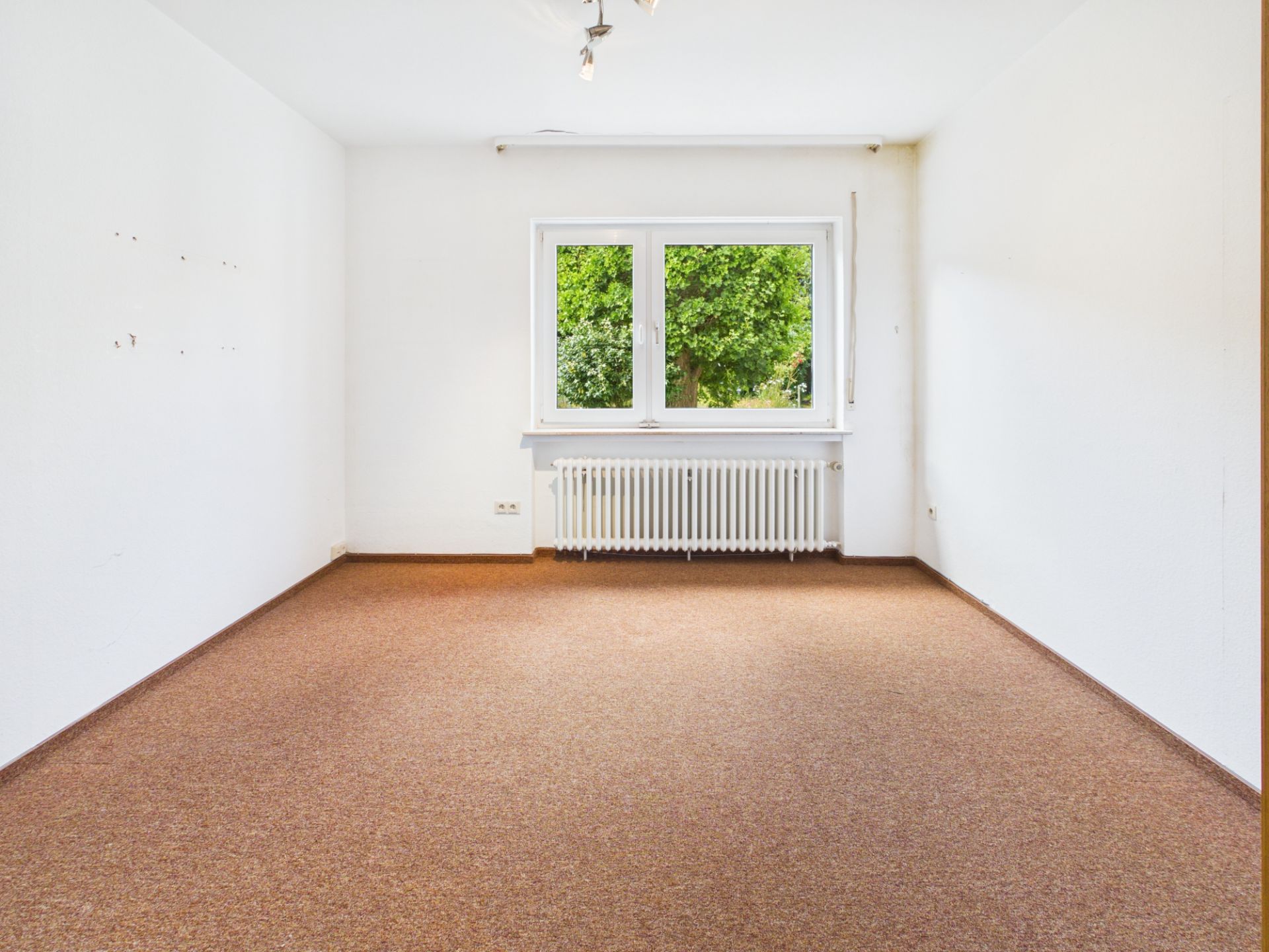 Leeres Zimmer mit braunem Teppich, weißer Wand, Fenster und Heizkörper.