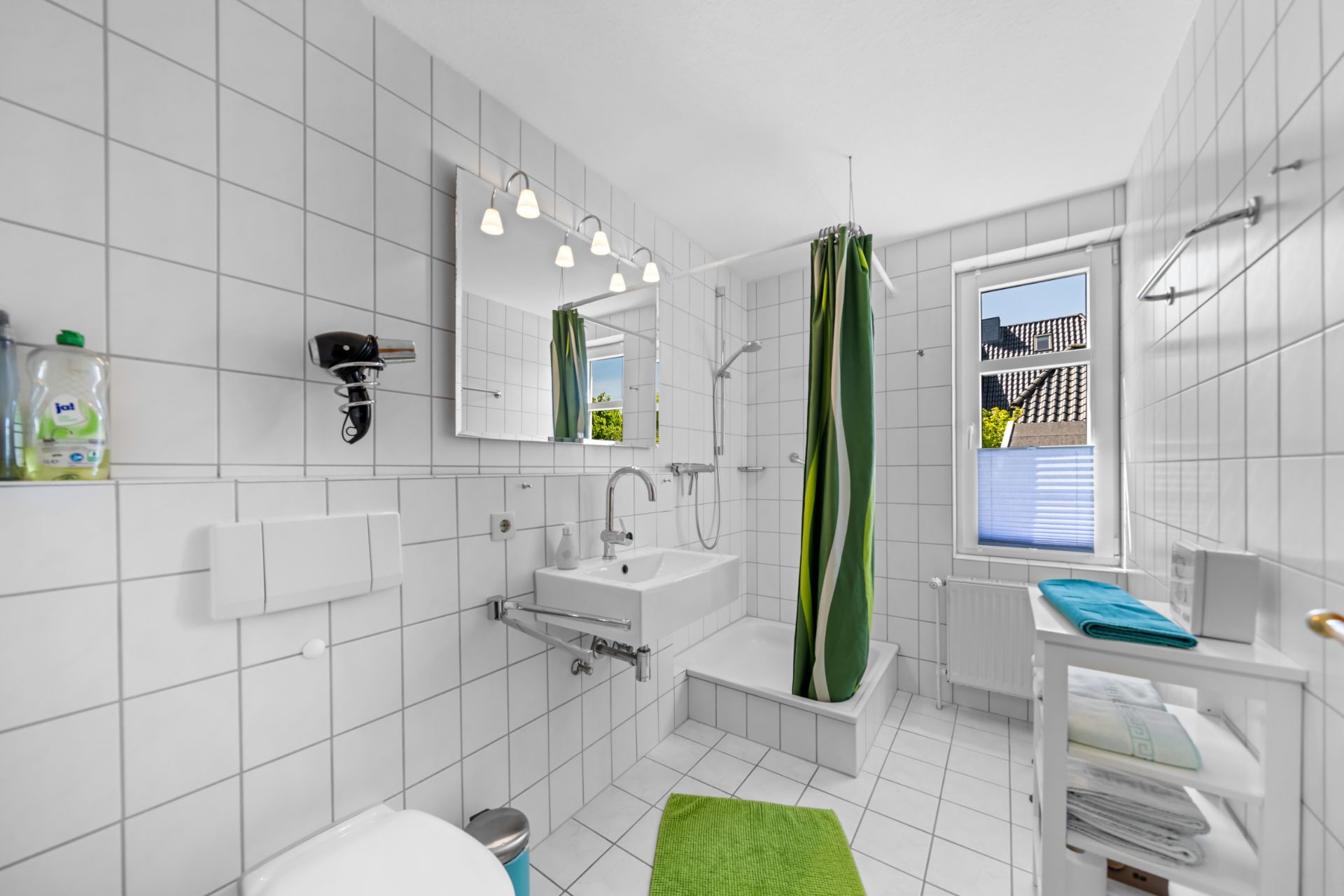 Helles Bad mit Dusche, grünem Duschvorhang, Spiegel, Waschbecken und Fenster.