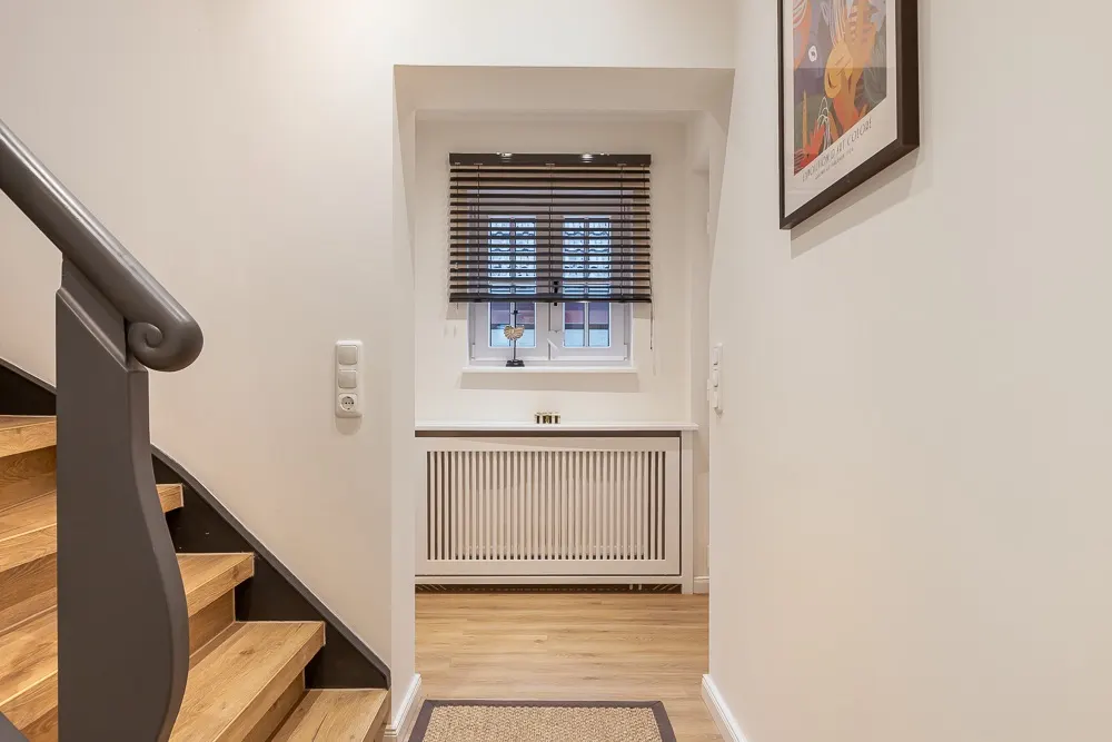 Flur mit Holztreppe, Fenster mit Jalousien und Heizkörper unter dem Fenster.